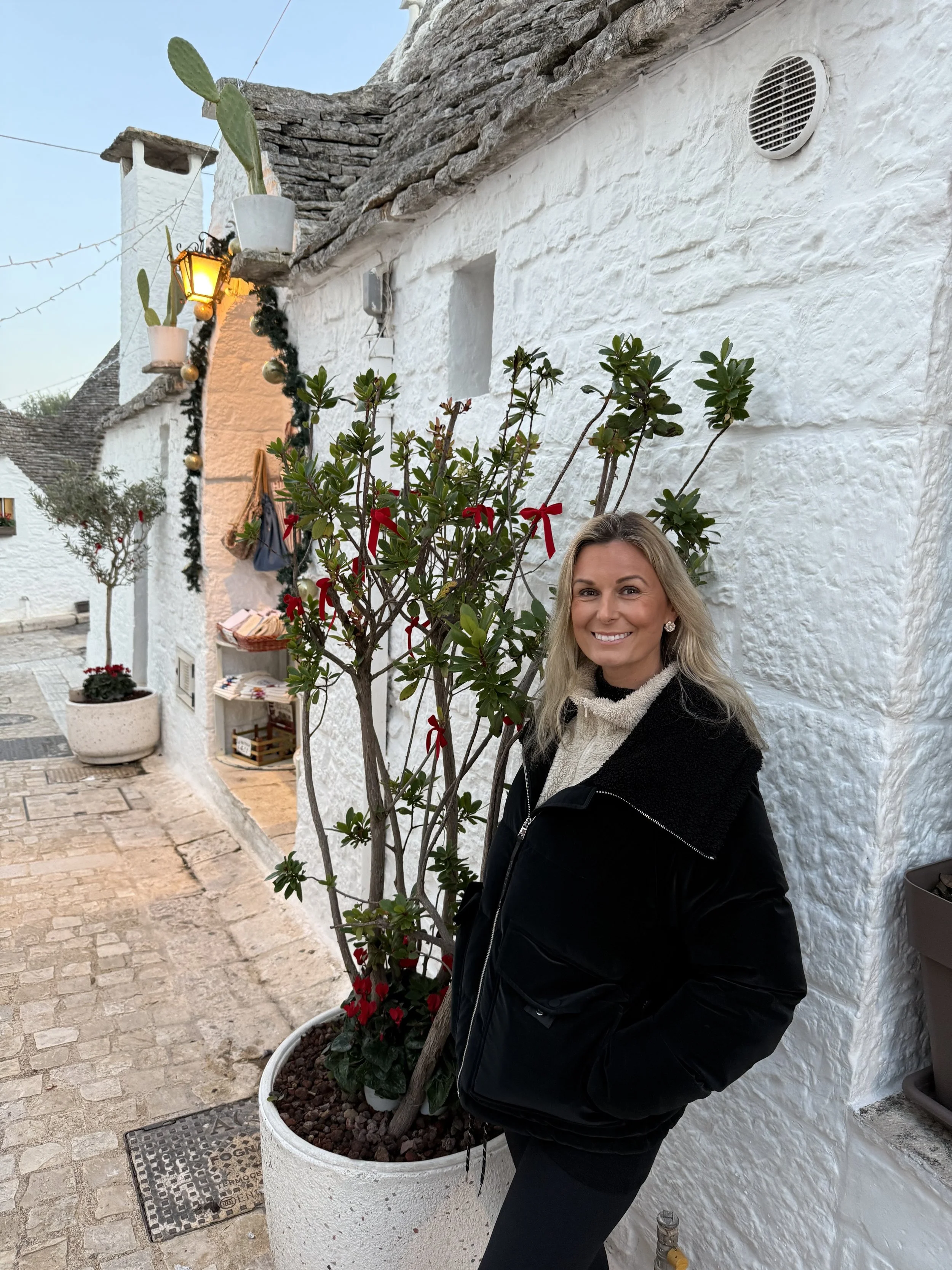 A smiling woman standing next to a potted plant with red flowers in front of a white stone wall. The setting appears to be an outdoor street or alley with cobblestone pavement and decorative pots, plants, and Christmas decorations.
