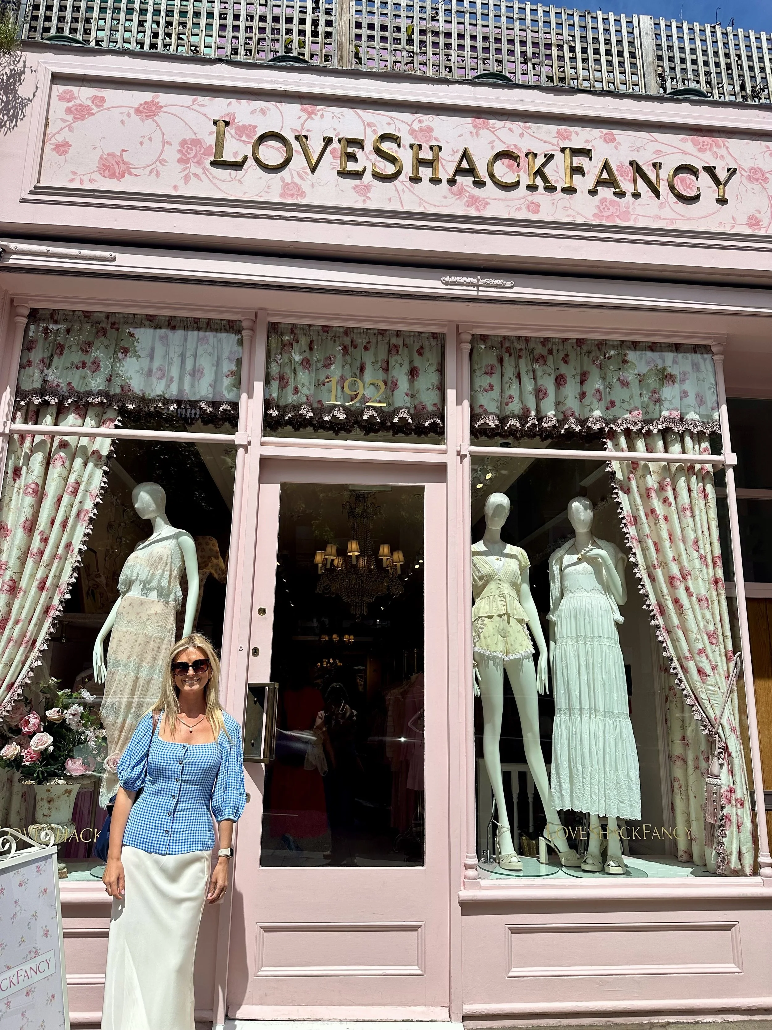 Woman with blonde hair wearing sunglasses, a blue checkered blouse, and a white skirt standing outside a boutique called LOVESHACKFANCY with pink exterior, floral curtains, and mannequins displaying dresses.