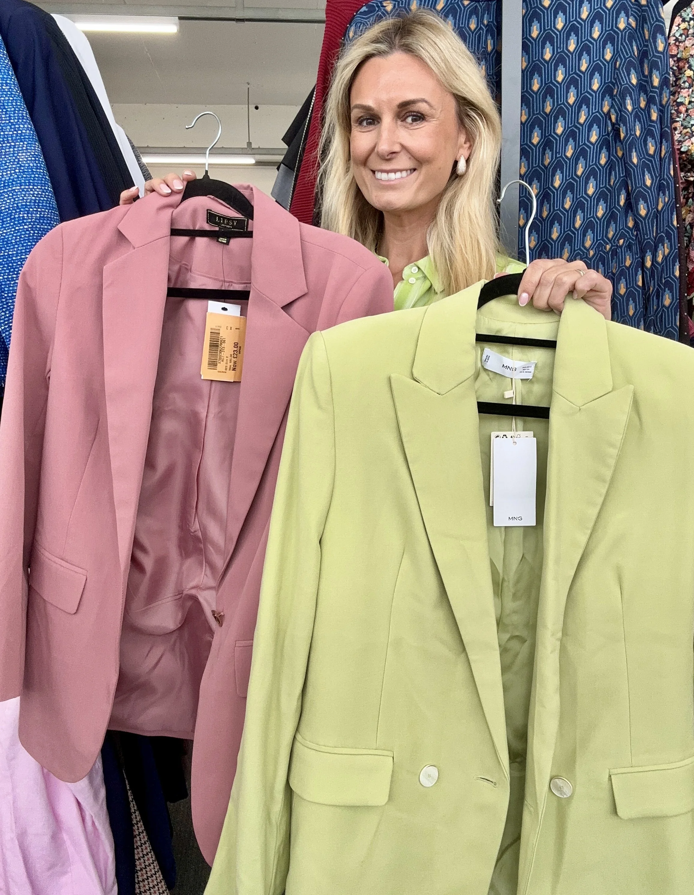 A woman smiling and holding two blazers, one pink and one yellow, on hangers inside a clothing store.