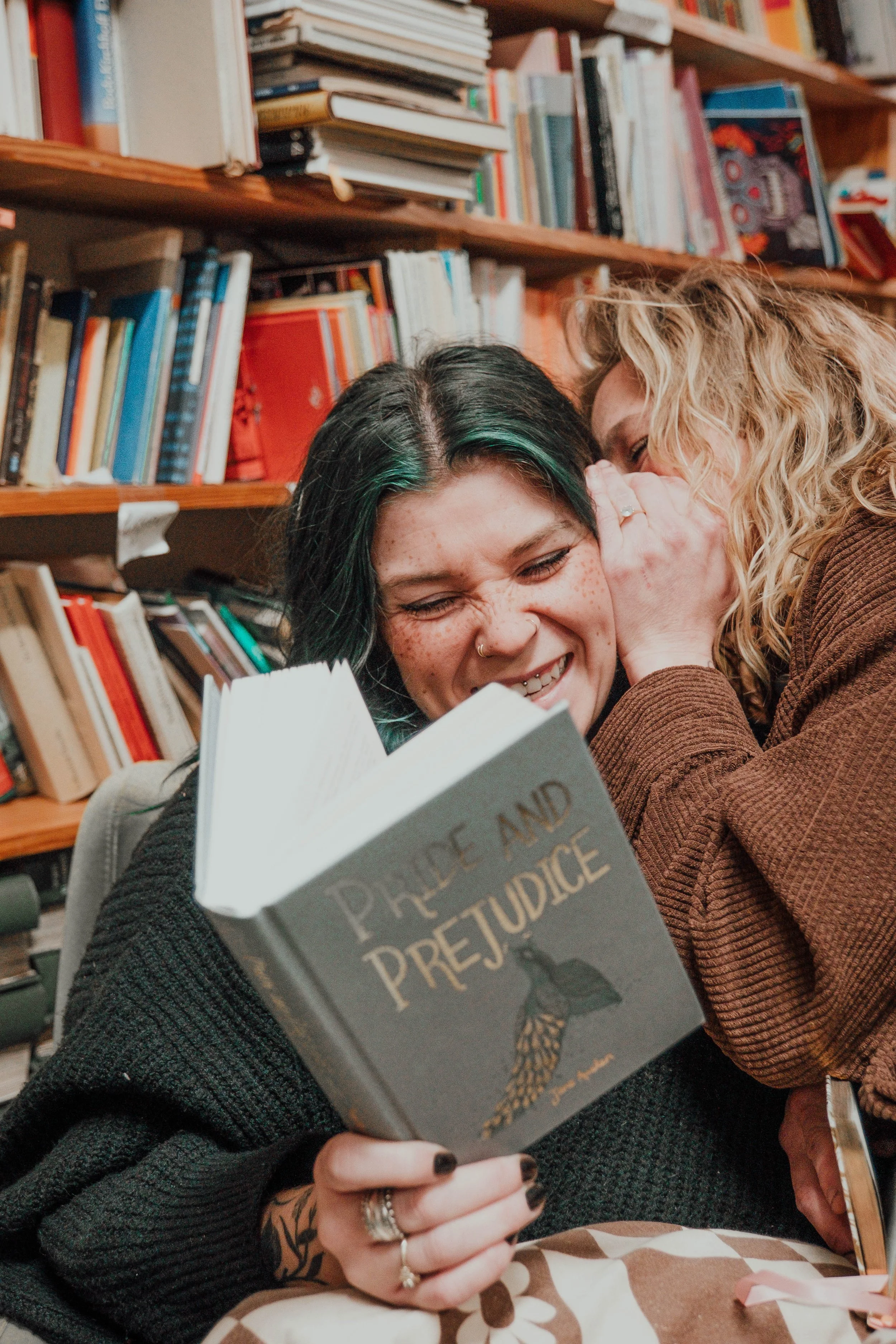 Two women sharing a secretive moment, with one woman whispering into the other's ear while the other looks at a book titled 'Pride and Prejudice', in a cozy library setting.