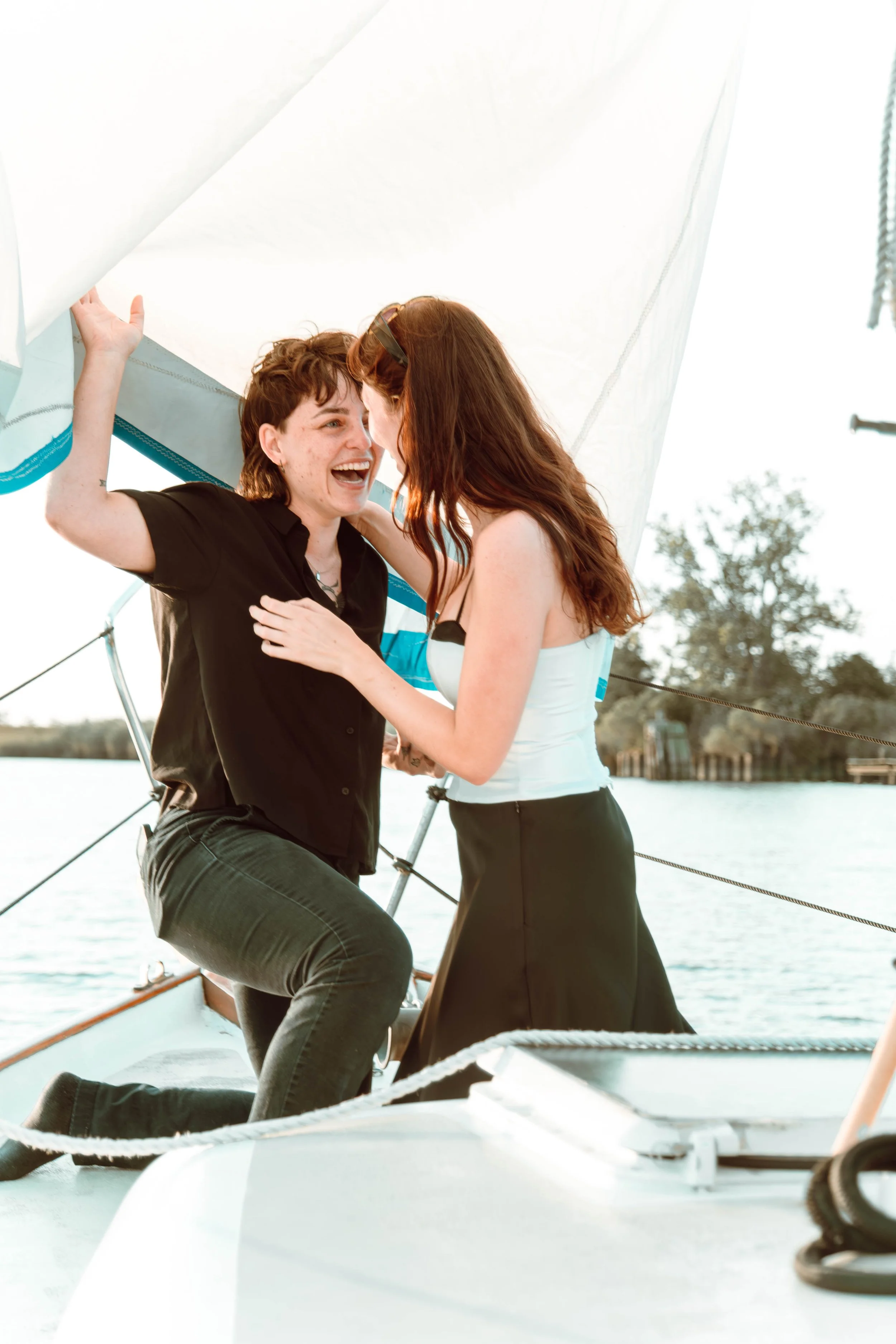 Two women on a sailboat laughing and embracing, with water and trees in the background.