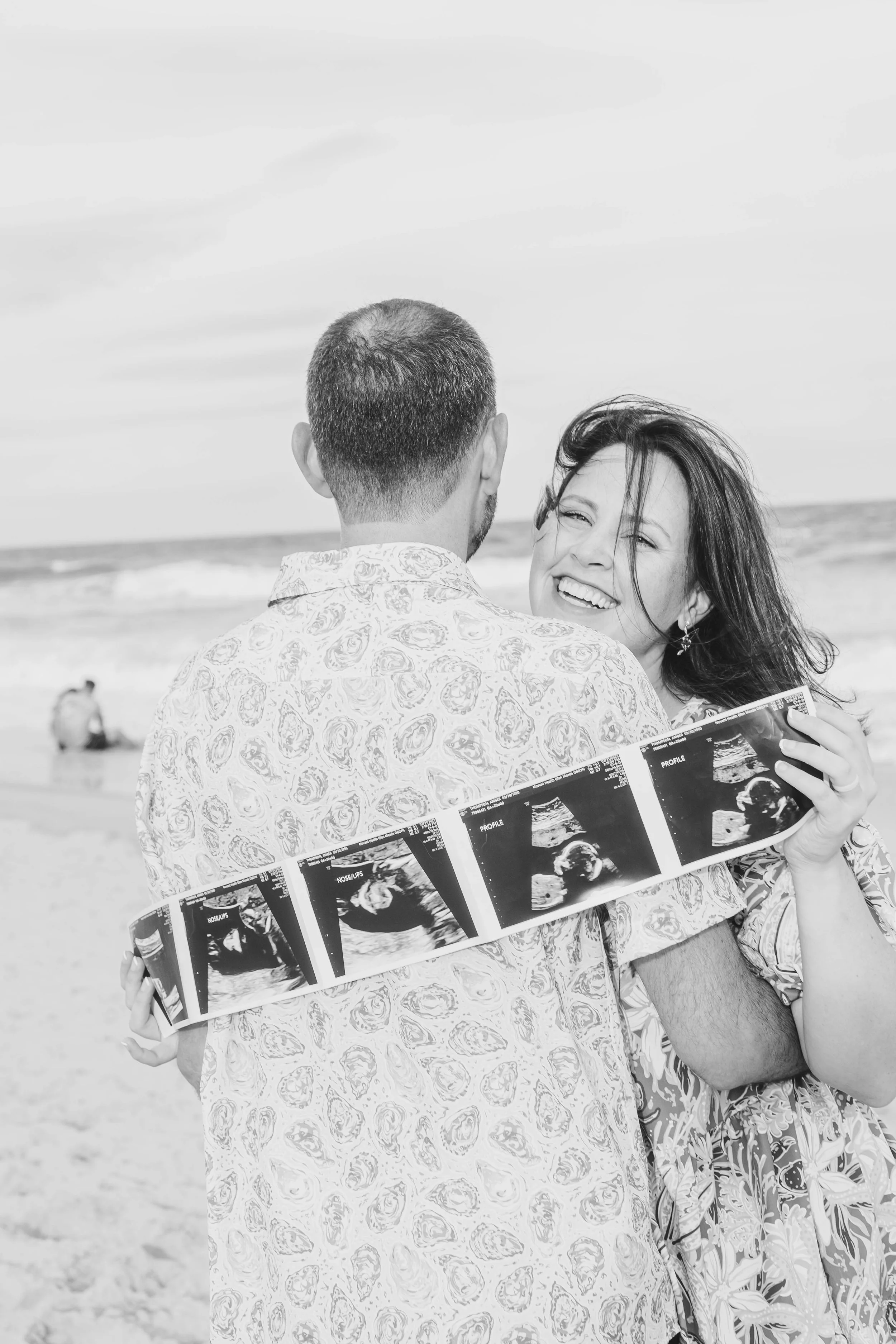 A man and woman at the beach holding ultrasound photos, with the woman smiling and the man facing away from the camera.