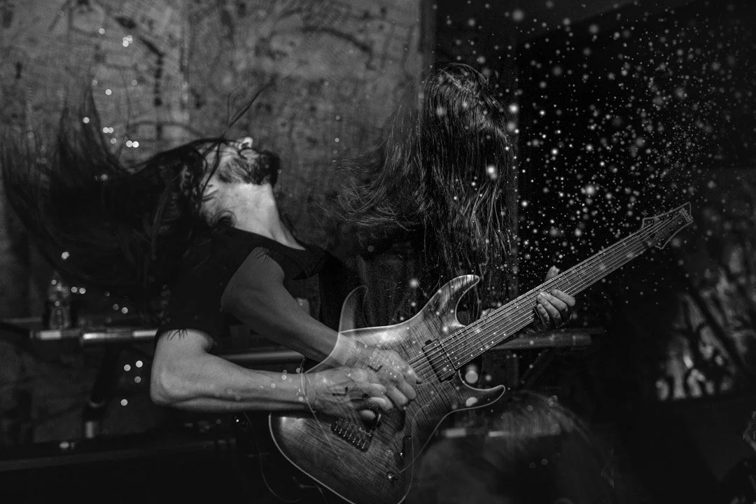 A guitarist performing with long hair, head tilted back, in front of a dark background with abstract light or snow effects.