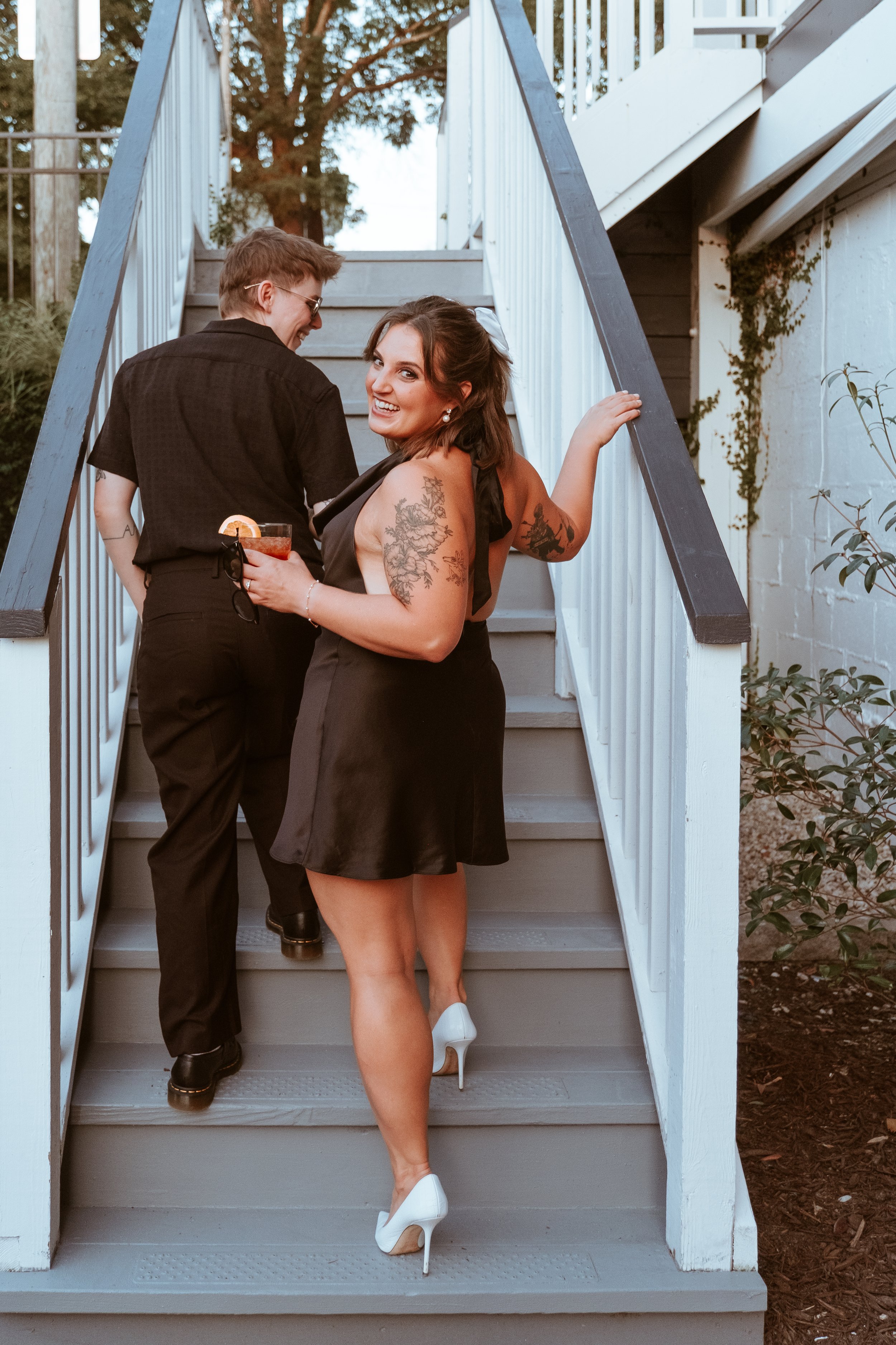 A woman wearing a black dress and white high heels smiling at the camera while holding a cocktail, with a man in black attire talking to someone off-camera on the stairs outside, with trees and greenery in the background.