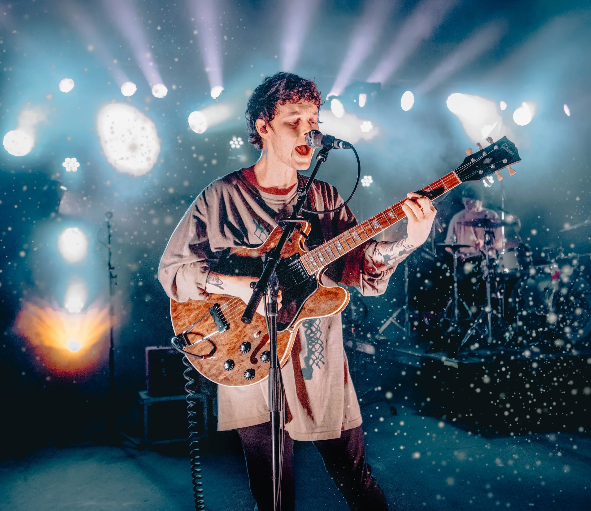 Musician singing and playing guitar during a concert with stage lights and background performer.