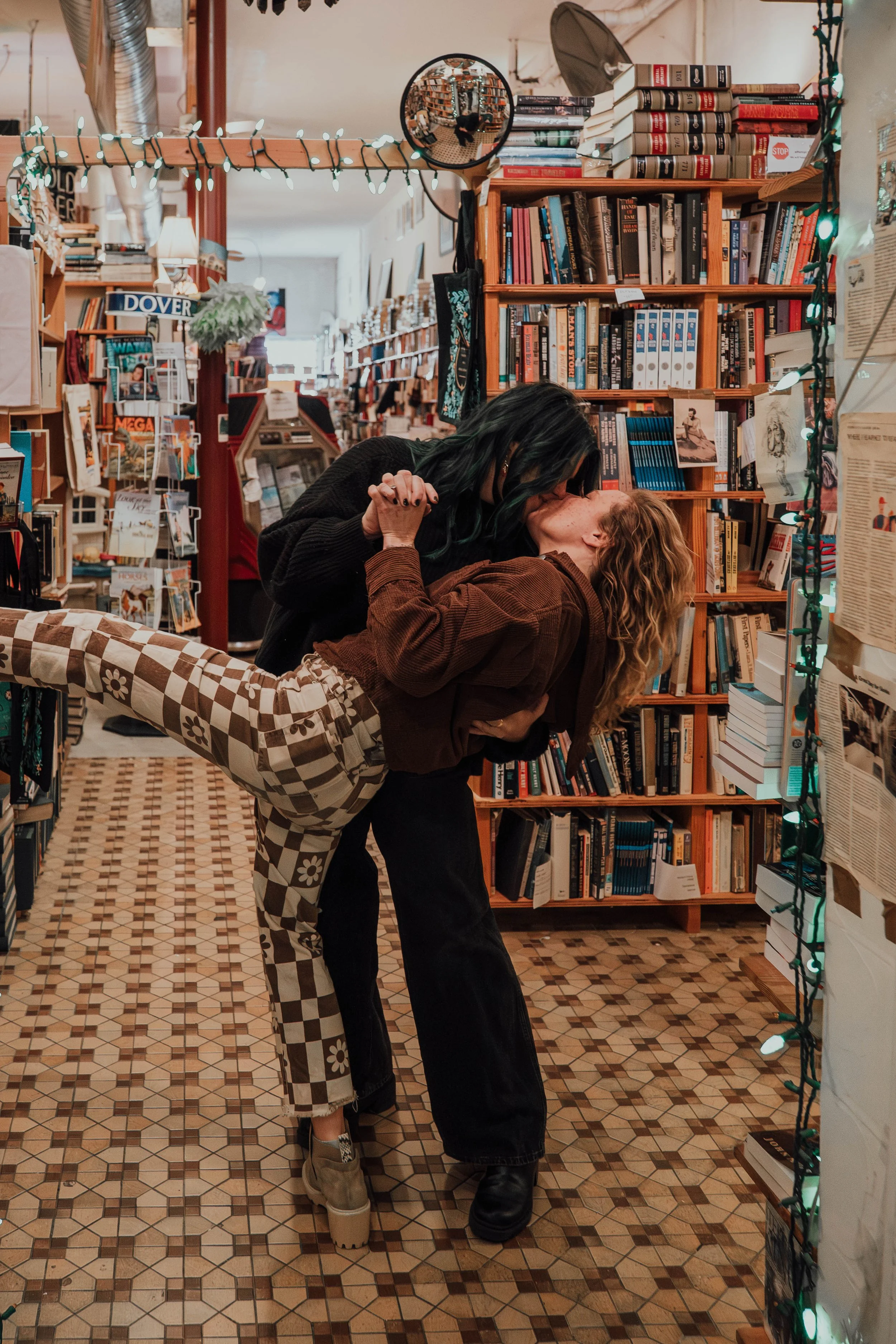 Two women kiss in a bookstore, one leaning back with an arm extended, the other bending forward. Bookshelves and decorative lights are visible in the background.