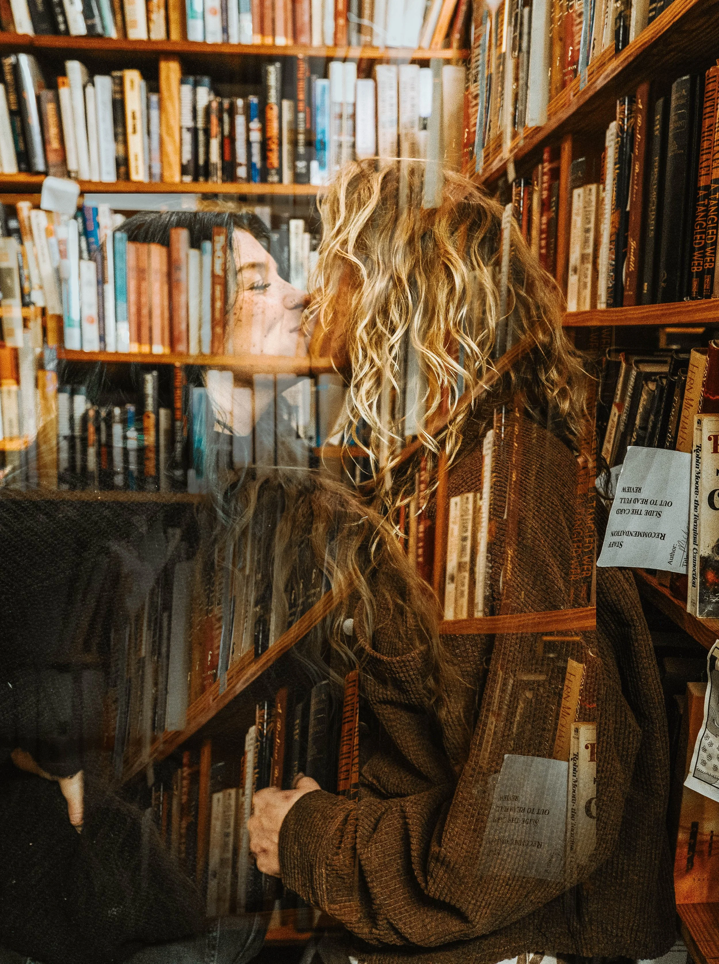 Two women with long, curly blonde hair are close to each other, separated by a glass shelf in a bookstore. They appear to be leaning in and possibly about to kiss, amidst shelves filled with books.