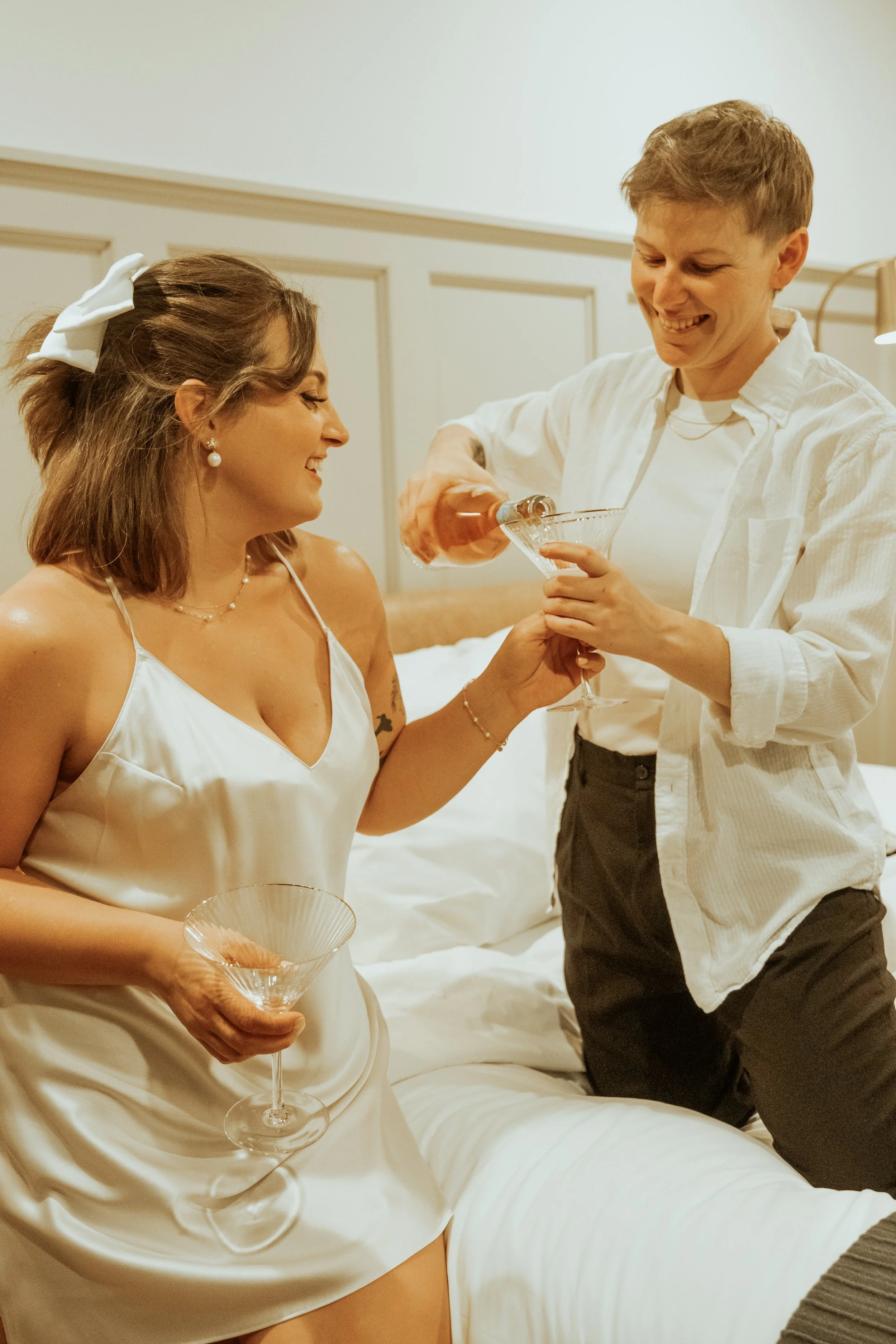A woman wearing a white satin slip dress holding a champagne glass while a man is pouring champagne into her glass, both smiling and celebrating in a cozy bedroom.