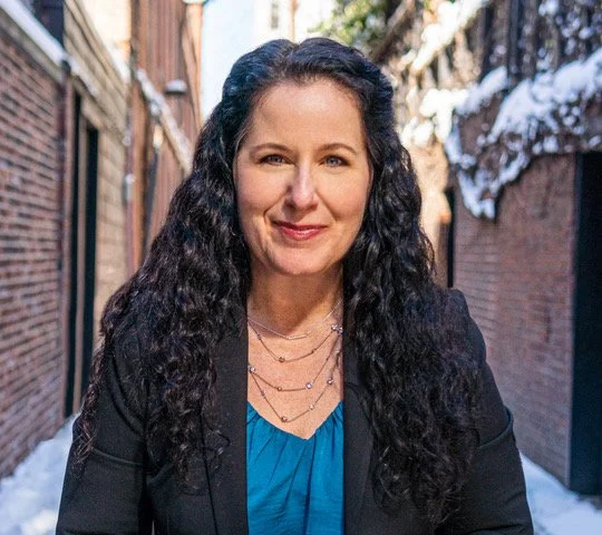 A woman with long curly dark hair smiling outdoors in a narrow alleyway with snow on the ground.