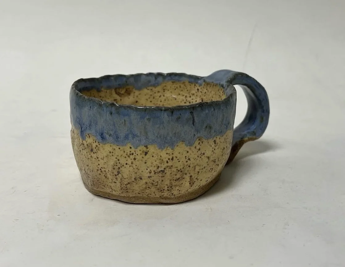A small handmade ceramic mug with a beige speckled body and a blue rim and handle.