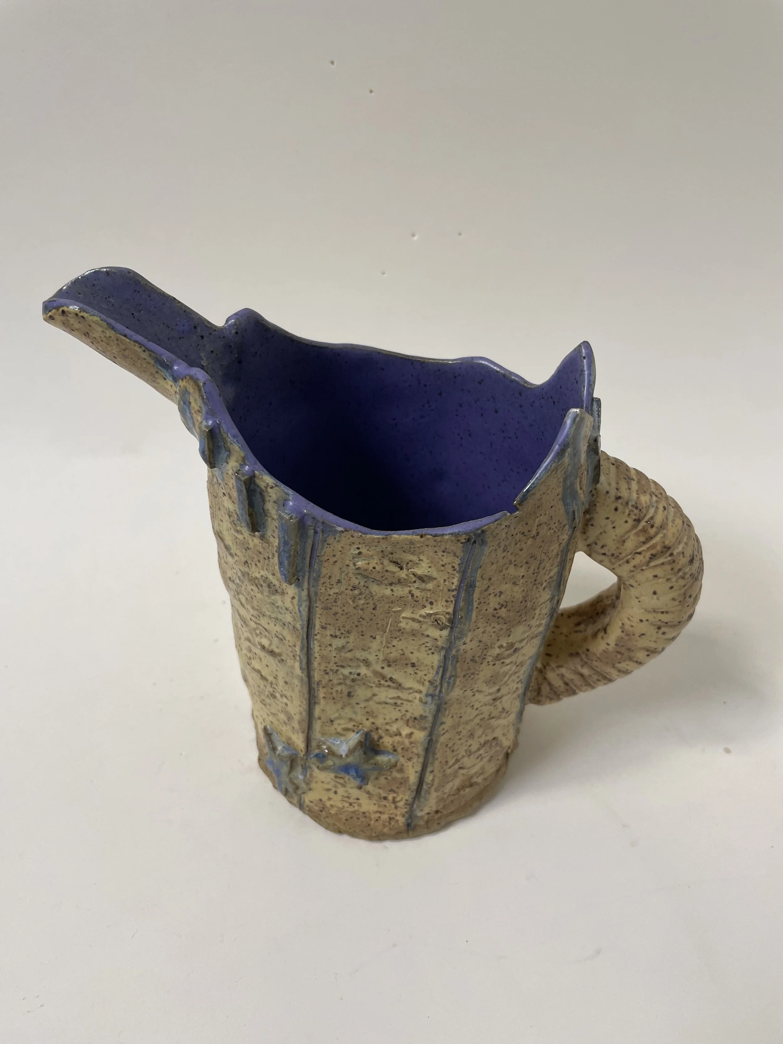 A small ceramic pitcher with a handle and a long lip, with a textured beige exterior decorated with blue stars and lines, and blue glaze inside.
