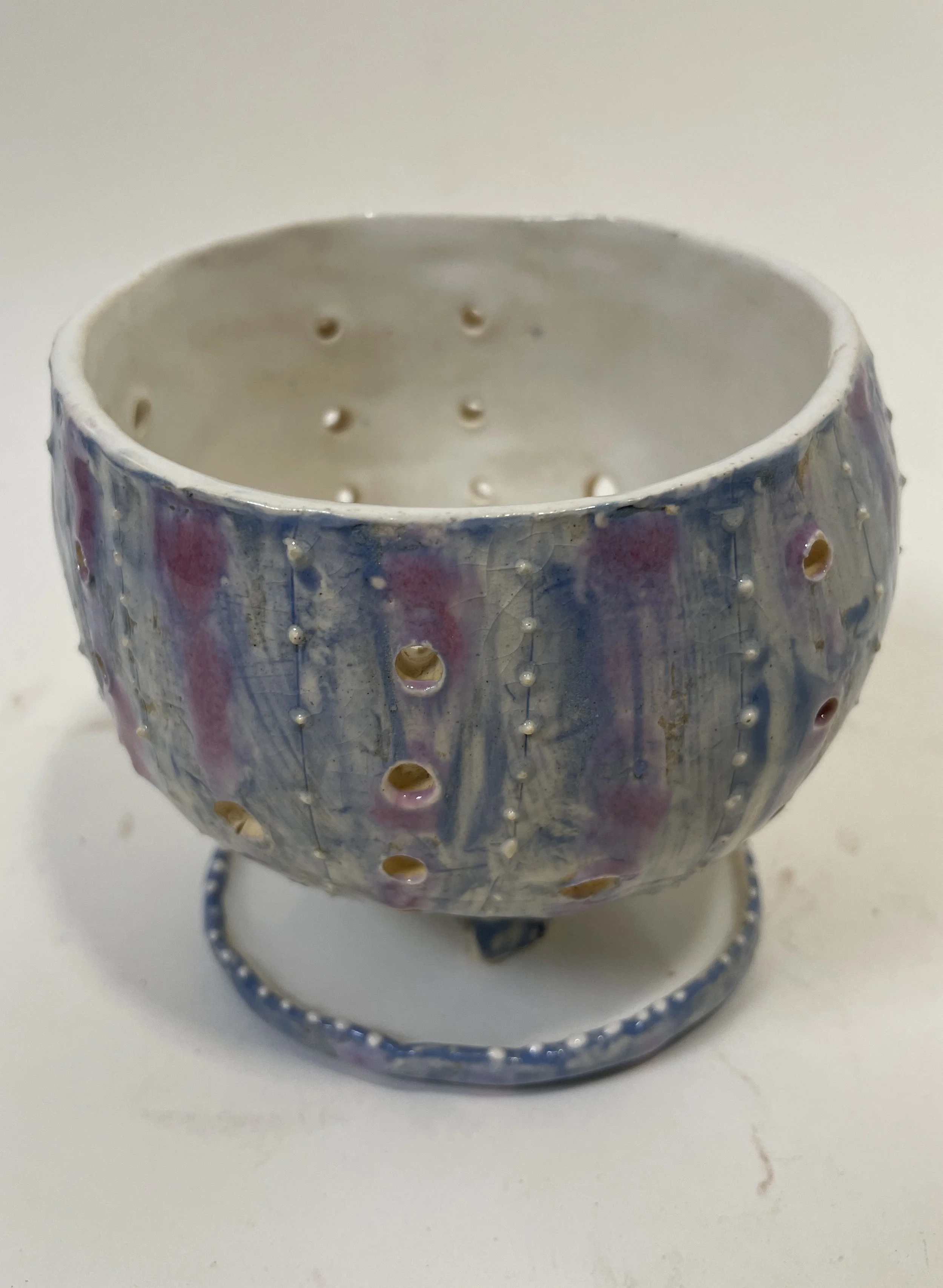 A small ceramic bowl with decorative holes in it, resting on a circular base, glazed in pale blue, pink, and purple.