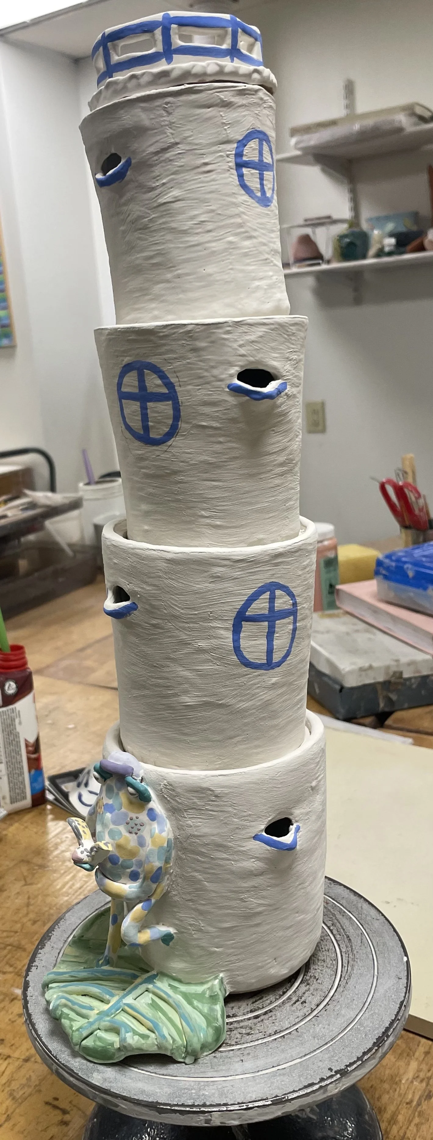 A tall, three-tiered ceramic sculpture of a lighthouse with windows and a seahorse at the base, all painted in soft pastel colors, on a pottery workshop table.