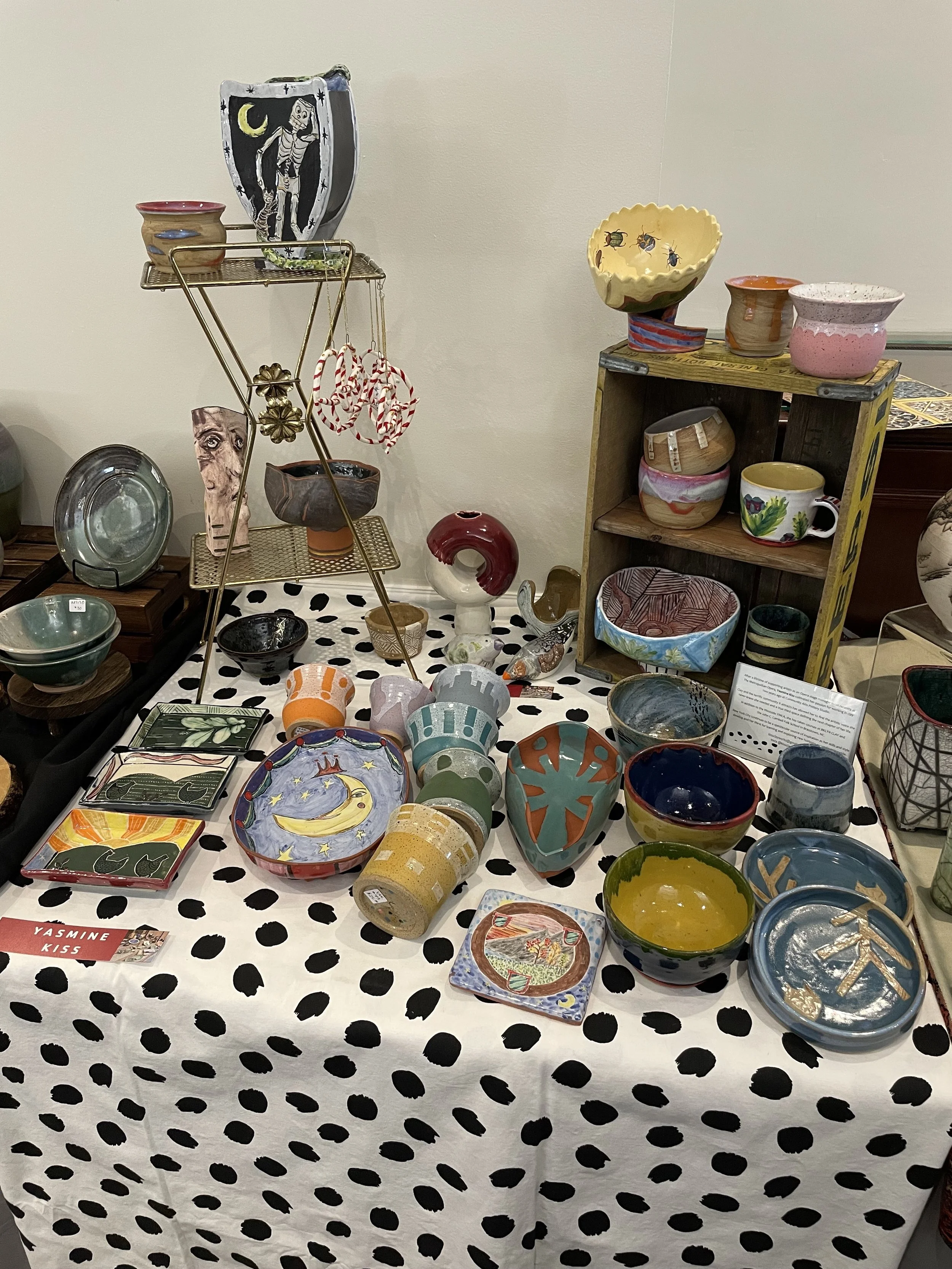 Display of colorful pottery and ceramic bowls on a table with a black and white polka dot tablecloth at a craft or art fair.