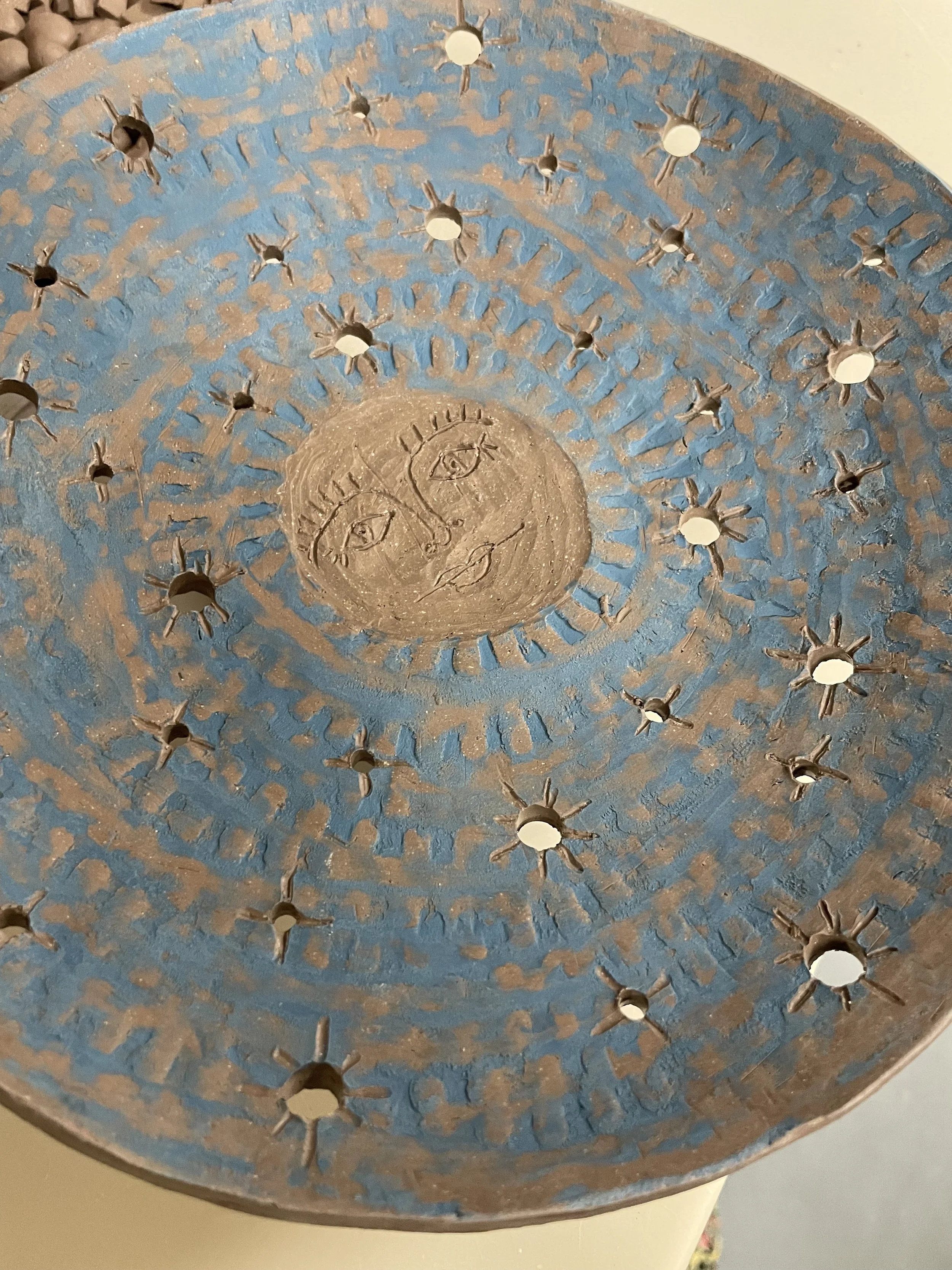 A blue and terracotta-colored ceramic dish with decorative holes, featuring a moon with a face in the center.