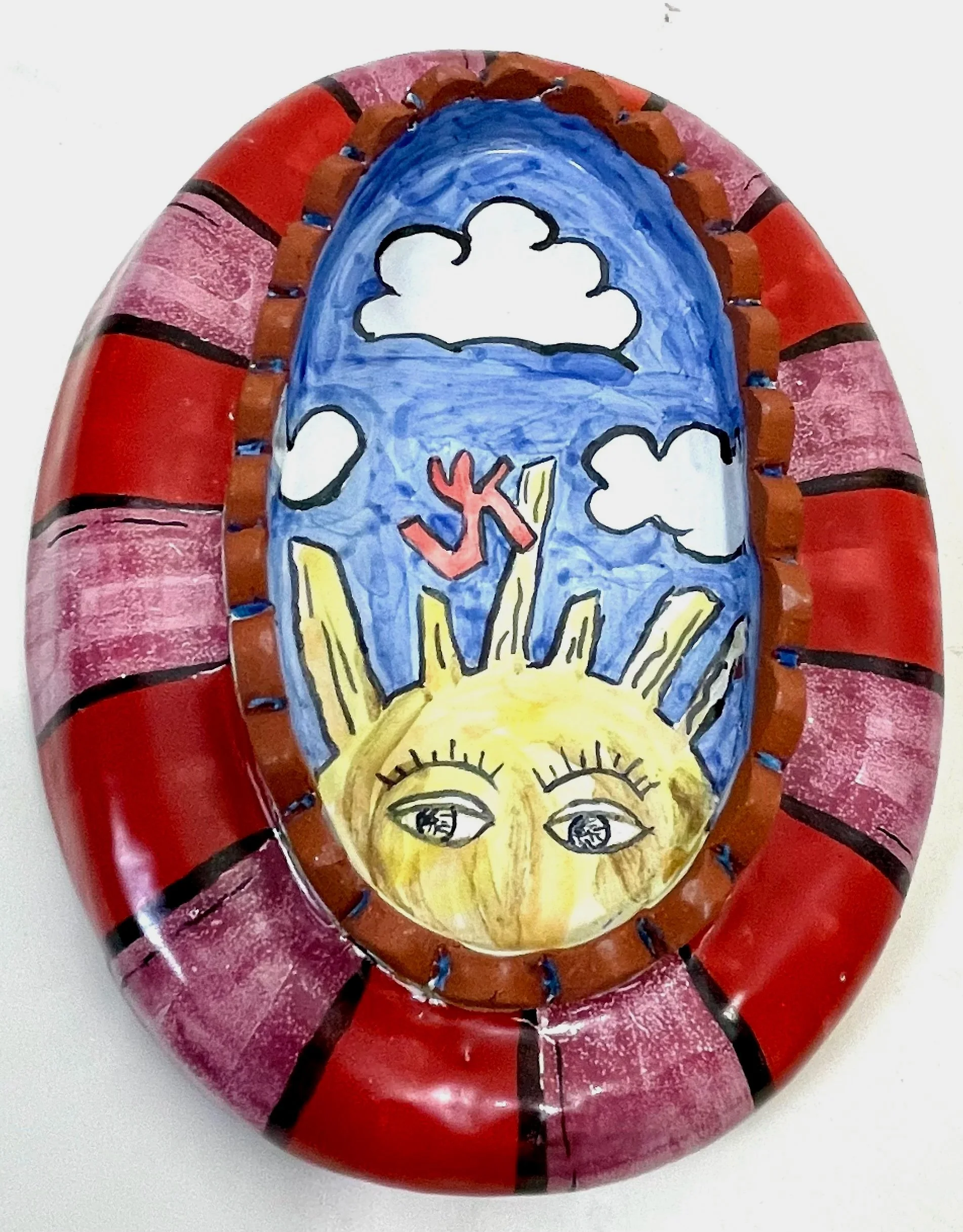 Decorative oval ceramics piece with a hand-painted scene of a yellow sun with eyes, mountains, clouds, and birds, surrounded by a red and pink striped border.