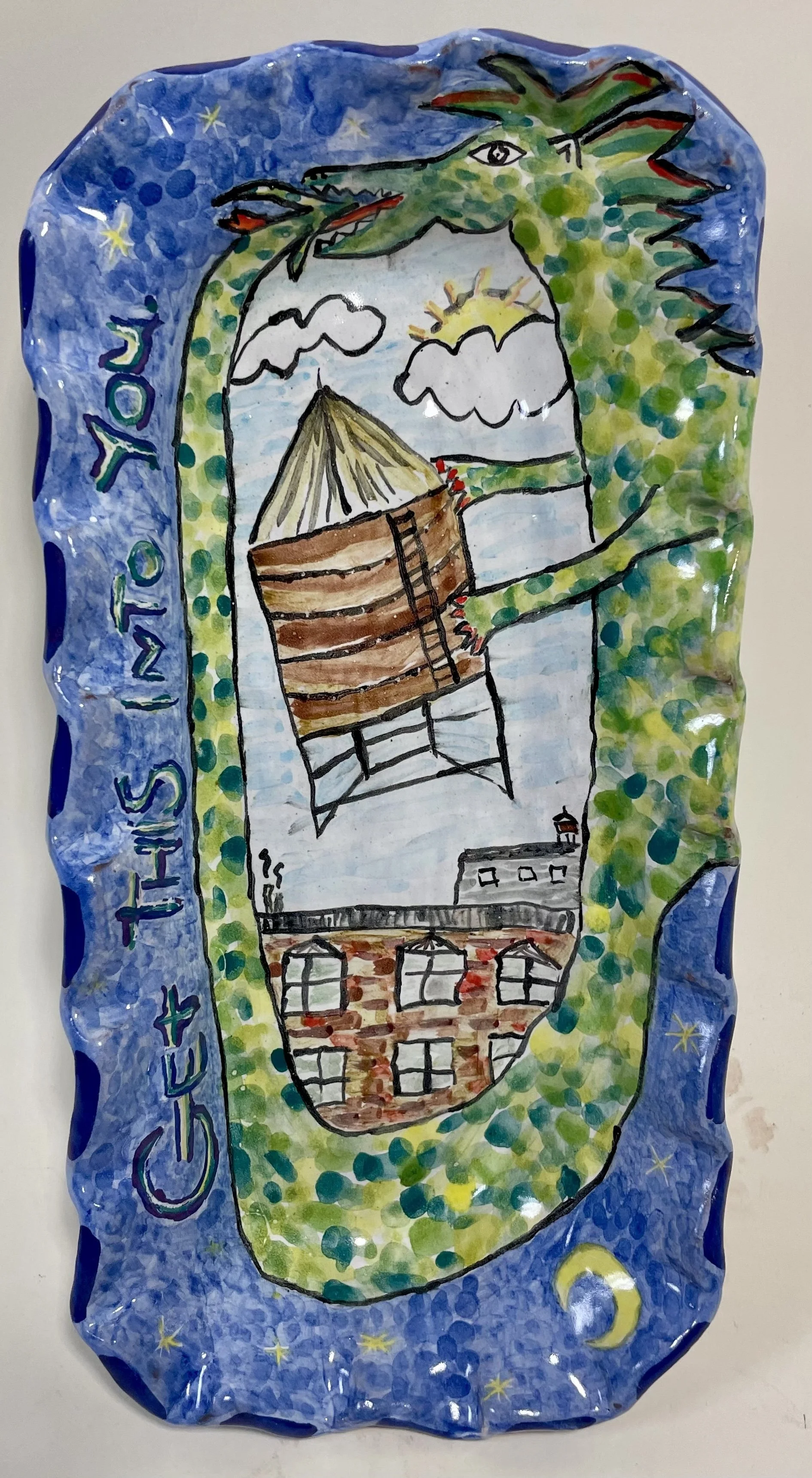 Colorful rectangular ceramic dish with a blue border and a painting of a green dragon encircling a brown building, lifting the top of the building off. Black text around the border reads "Get This Into You."