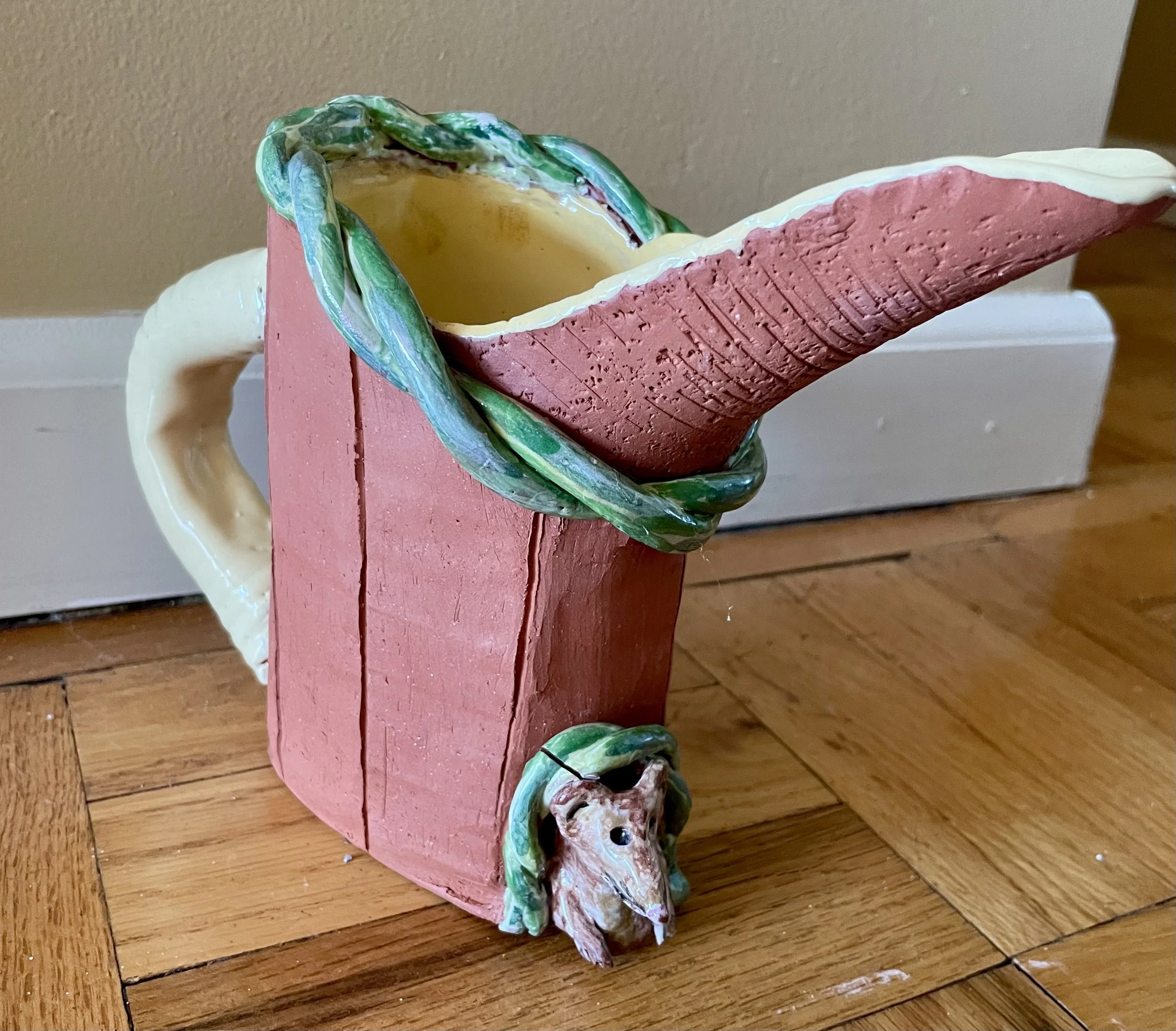 An unfired reddish-colored ceramic pitcher with a gray handle, green braiding around the lip, and a sculpted gray mouse peeking out of the bottom.