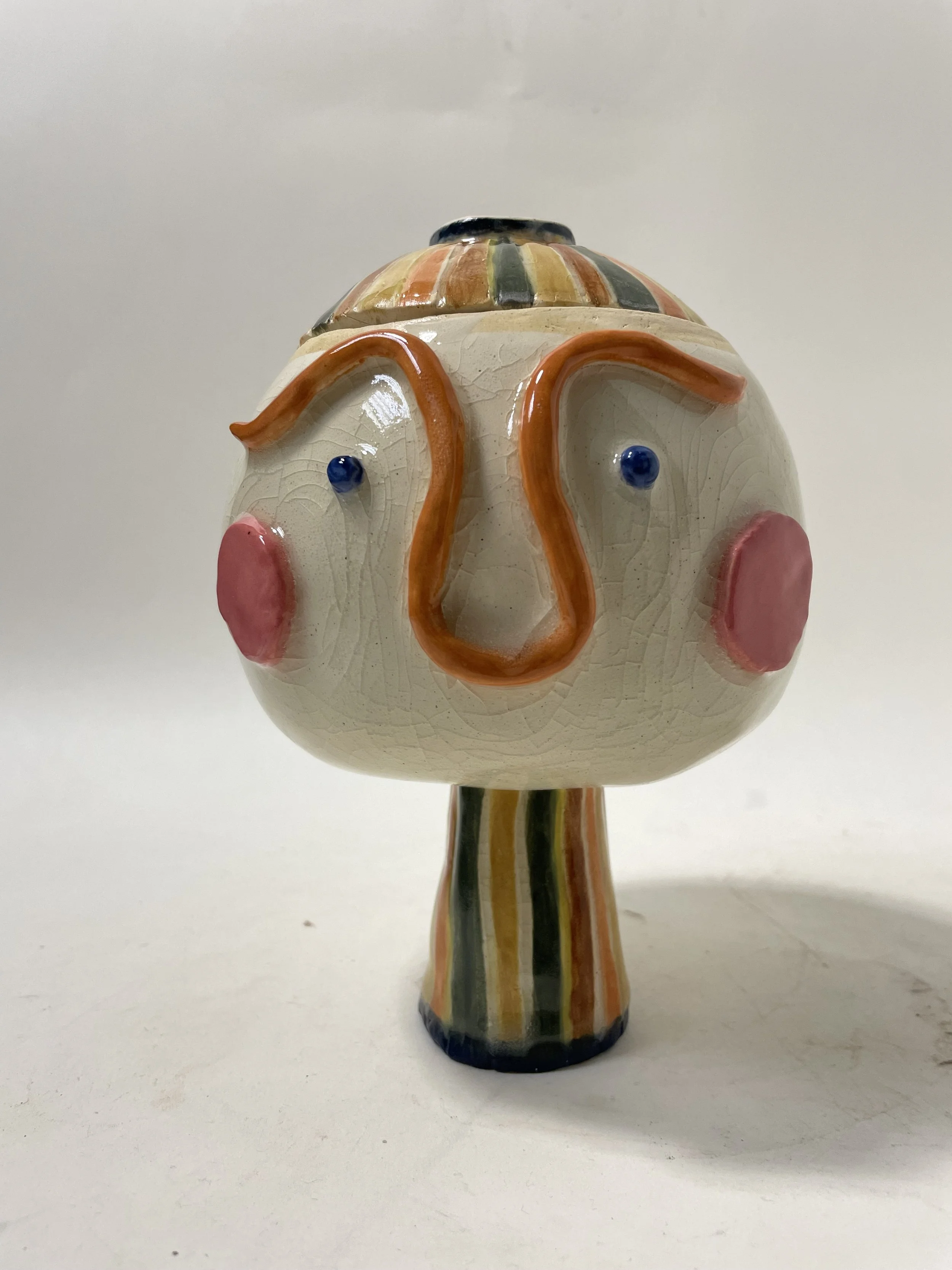 A colorful ceramic vase shaped like a face with exaggerated features including a long orange nose, pink cheeks, and small blue eyes, with a striped pattern on the top and base.