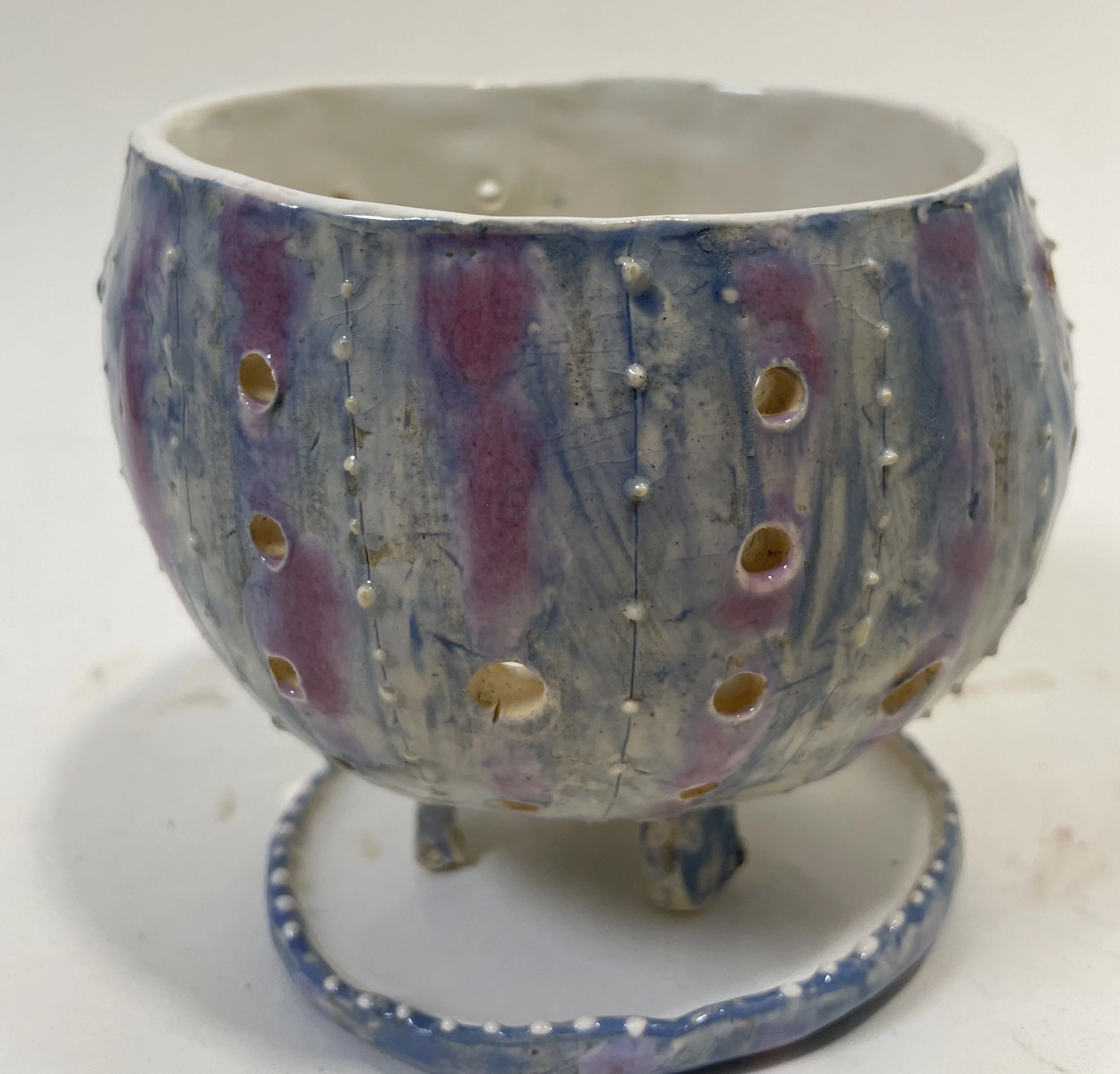 A small ceramic bowl with decorative holes in it, resting on a circular base, glazed in pale blue, pink, and purple.