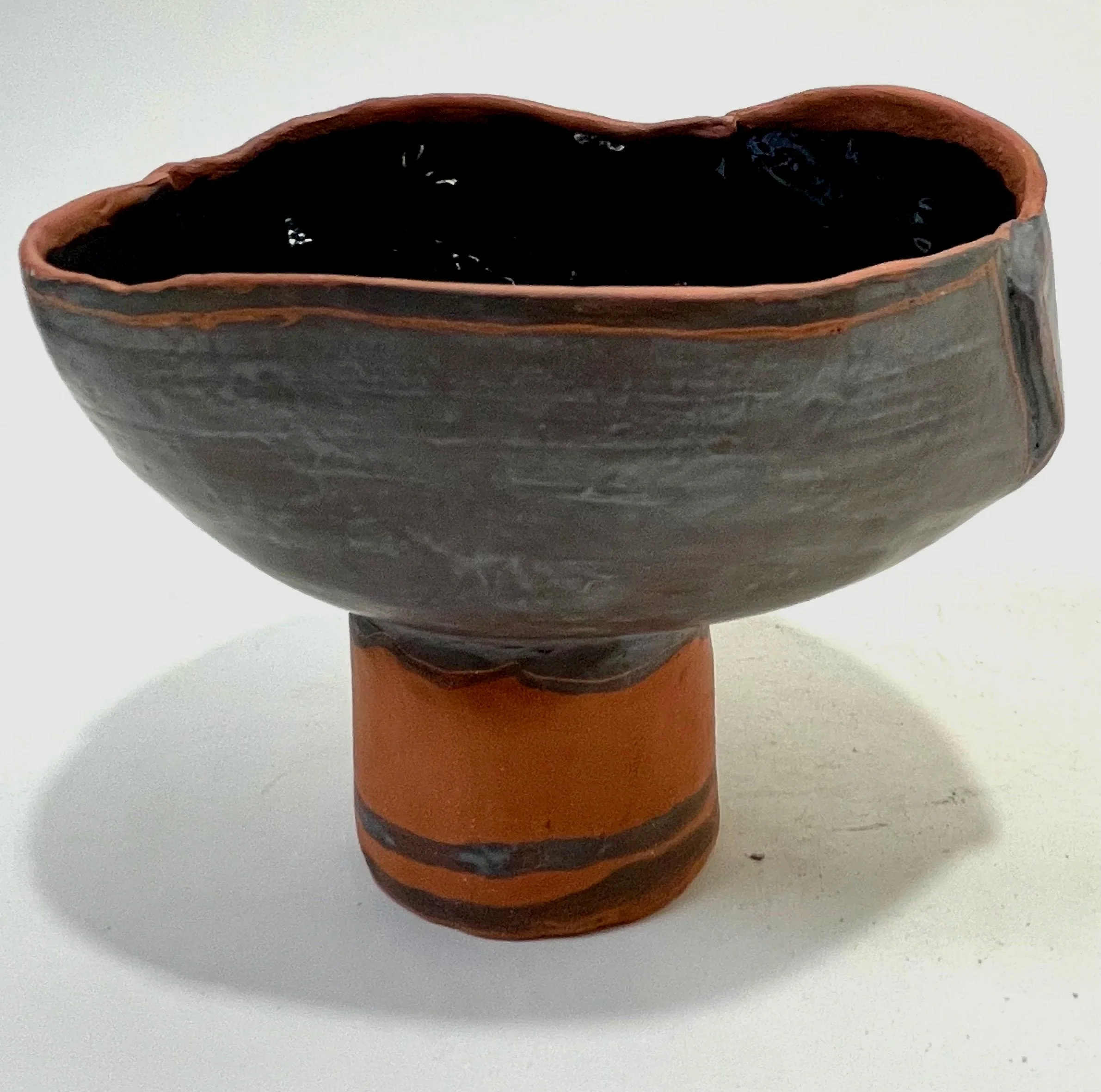 A ceramic vessel with a wide bowl on a pedestal base, glazed in dark gray and terracotta colors, with black glaze inside.