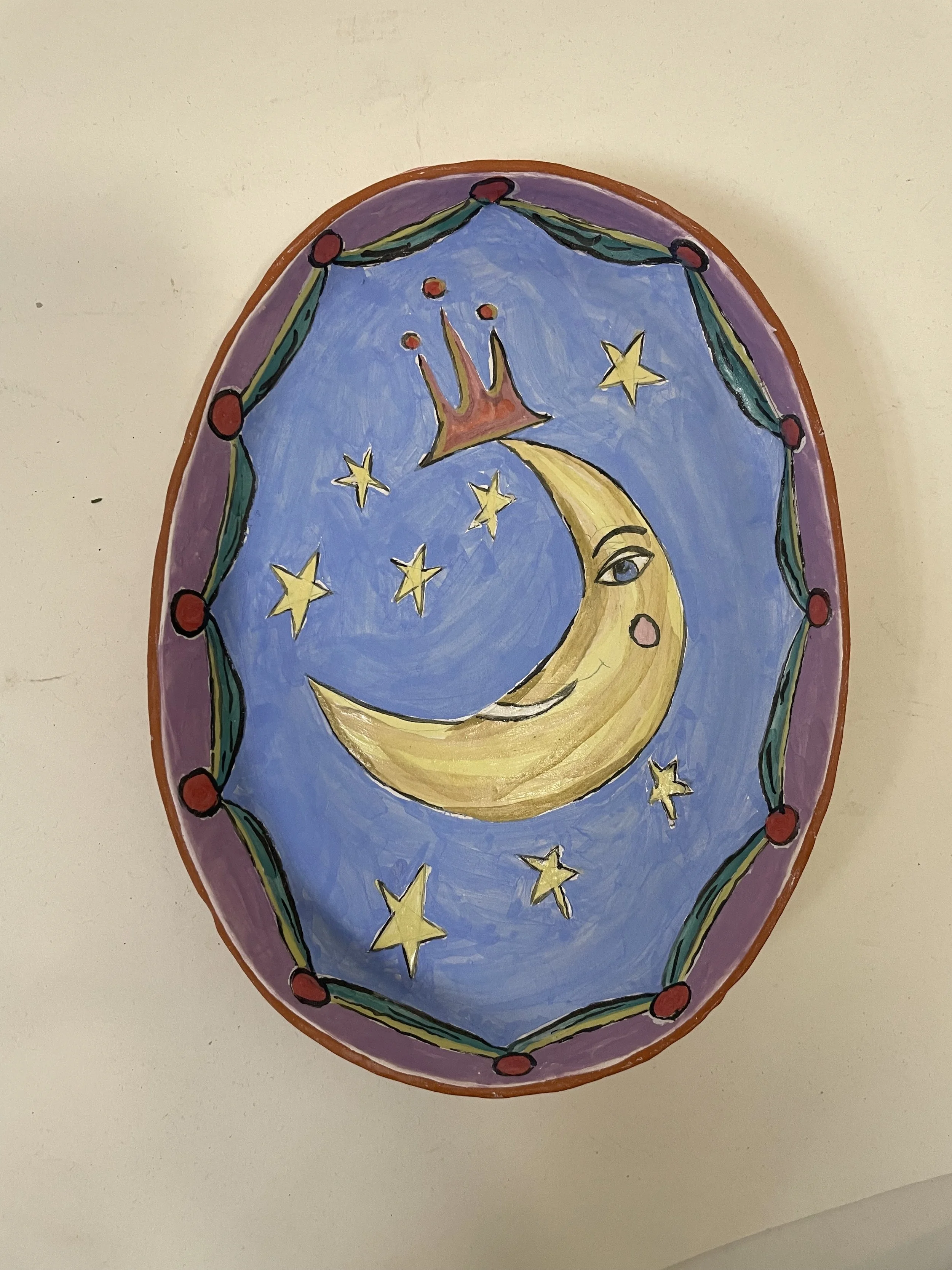 A colorful, round ceramic dish before kiln firing, depicting a pale blue night sky with yellow stars and a yellow crescent moon wearing a crown, bordered by a decorative purple and green pattern.