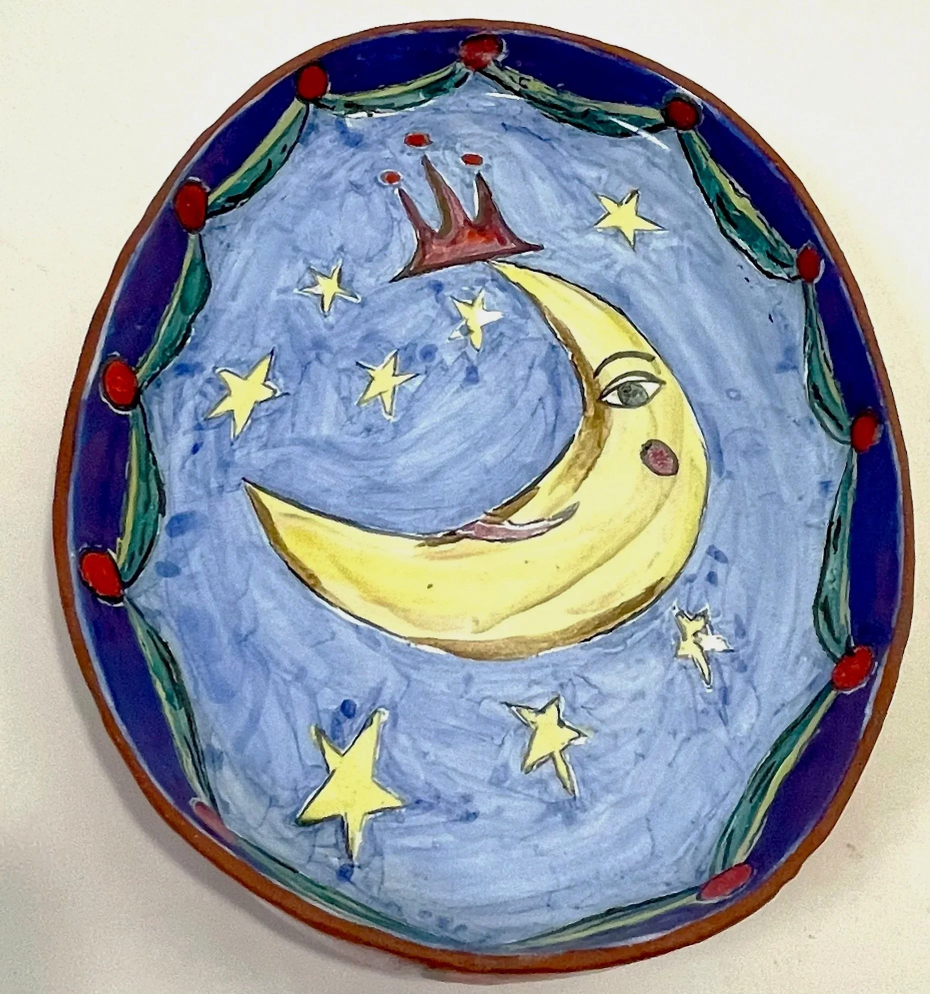 A colorful, round ceramic dish after kiln firing, depicting a pale blue night sky with yellow stars and a yellow crescent moon wearing a crown, bordered by a decorative purple and green pattern.