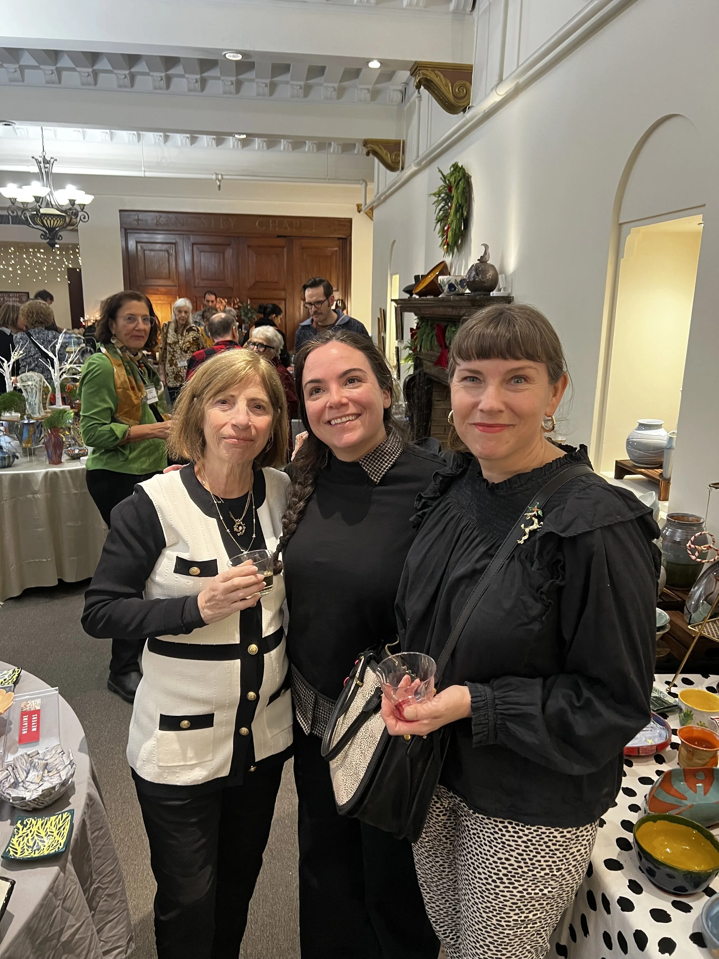 Three women standing together at a social event, smiling. One woman is holding a drink, another has a purse, and they are in a decorated room with tables and other people in the background.