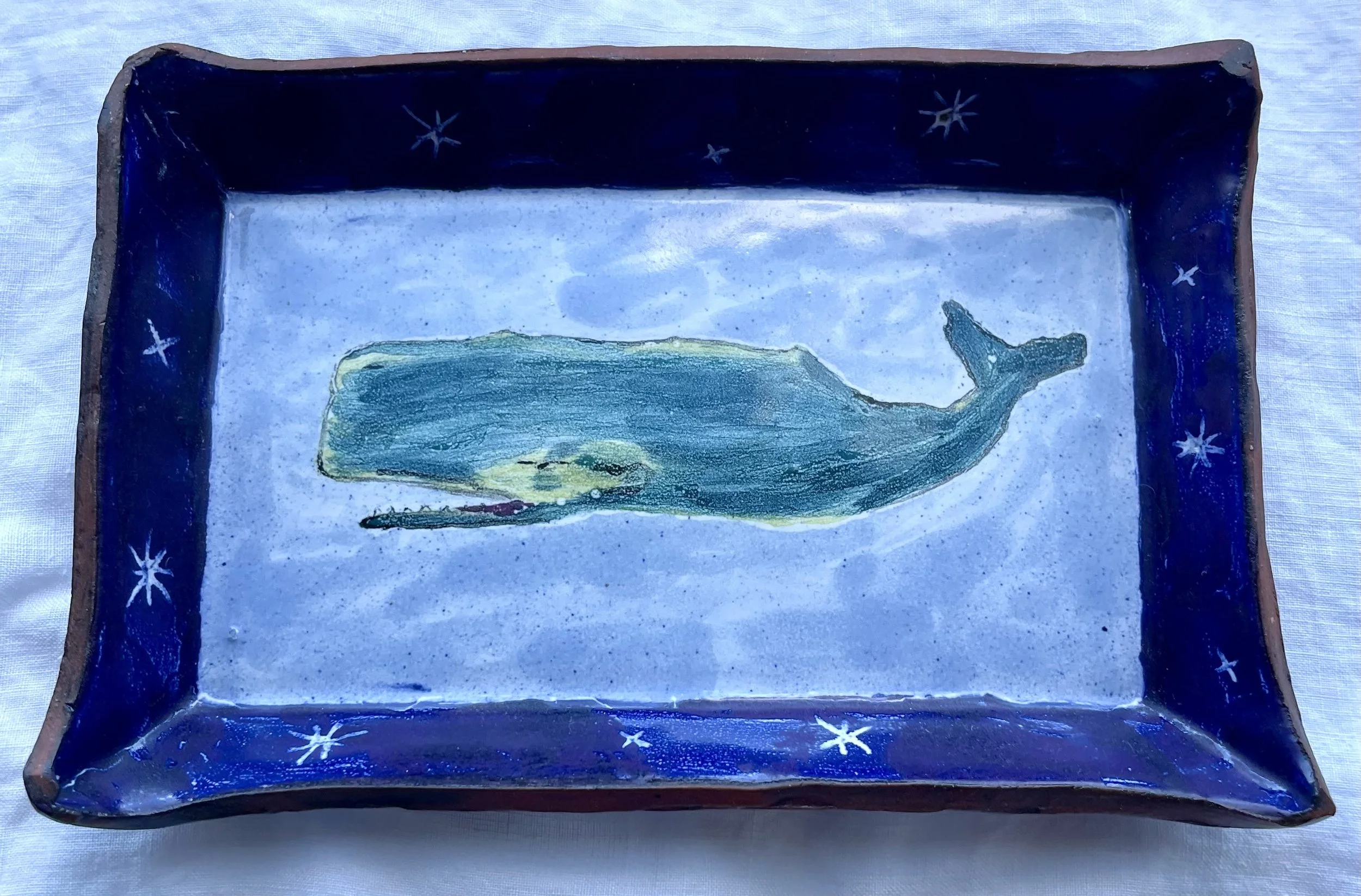 A rectangular ceramic tray with a dark blue border decorated with white stars, and a pale bue interior featuring a whale in shades of blue and green.