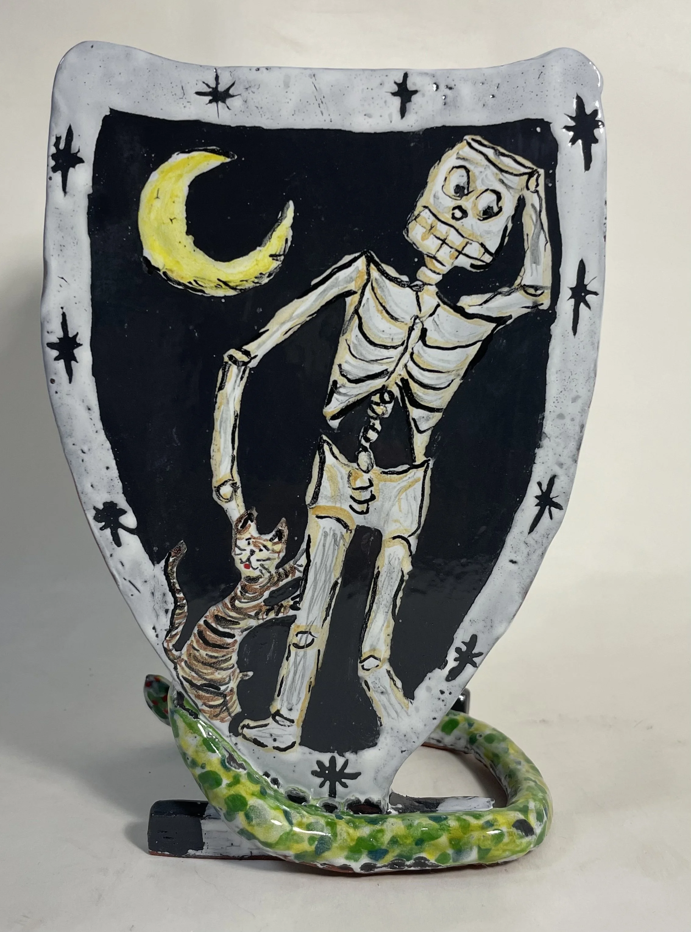 Decorative ceramic shield featuring a painted skeleton and a cat below it, with a yellow crescent moon in the top left corner and star patterns around the border.