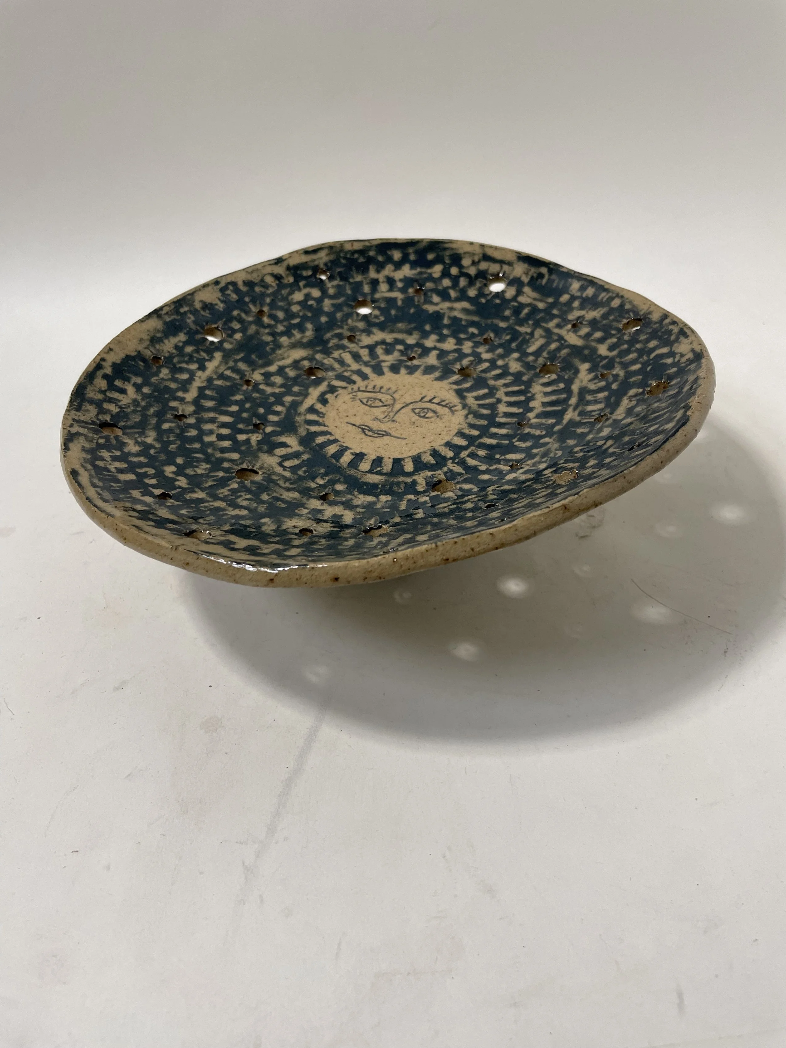 A blue-glazed ceramic dish with a moon face in the center and small decorative holes all over the surface.