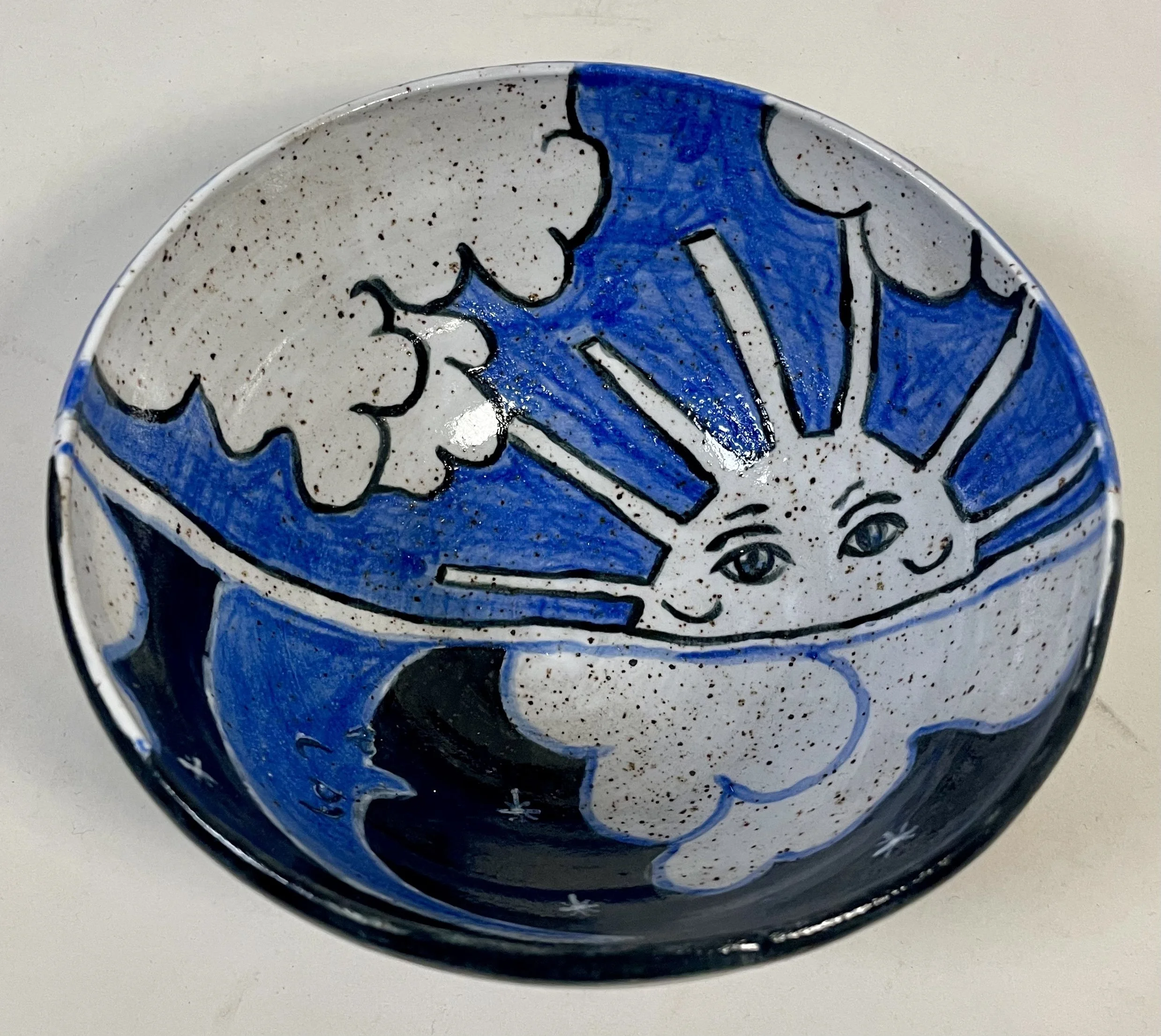 Inside of a decorative bowl featuring a smiling sun with rays, a moon with a face, and clouds, all painted in blue, black, and white.