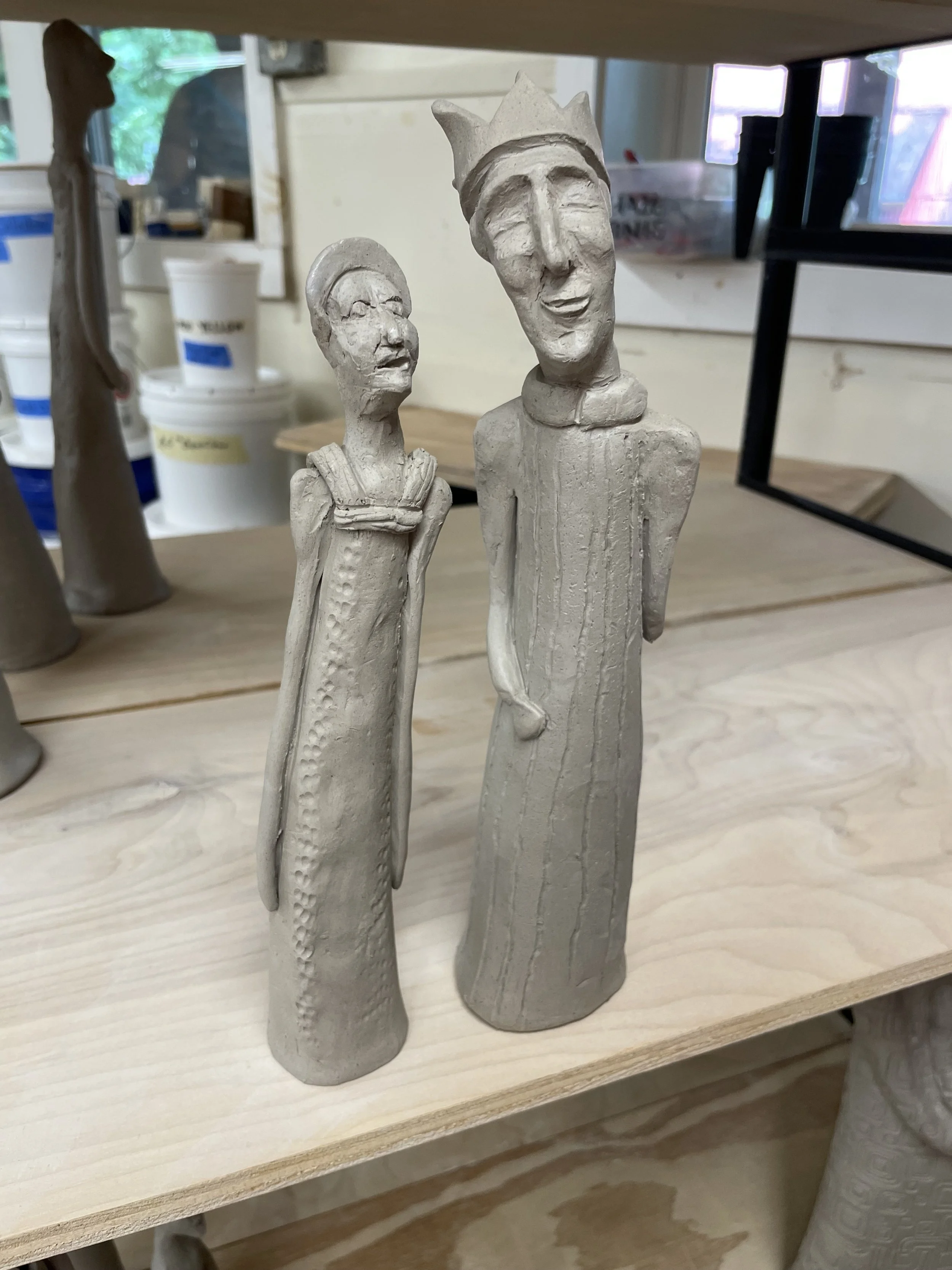 Two elongated ceramic sculptures of people, one wearing a crown and the other wearing glasses, waiting to be fired in the kiln.