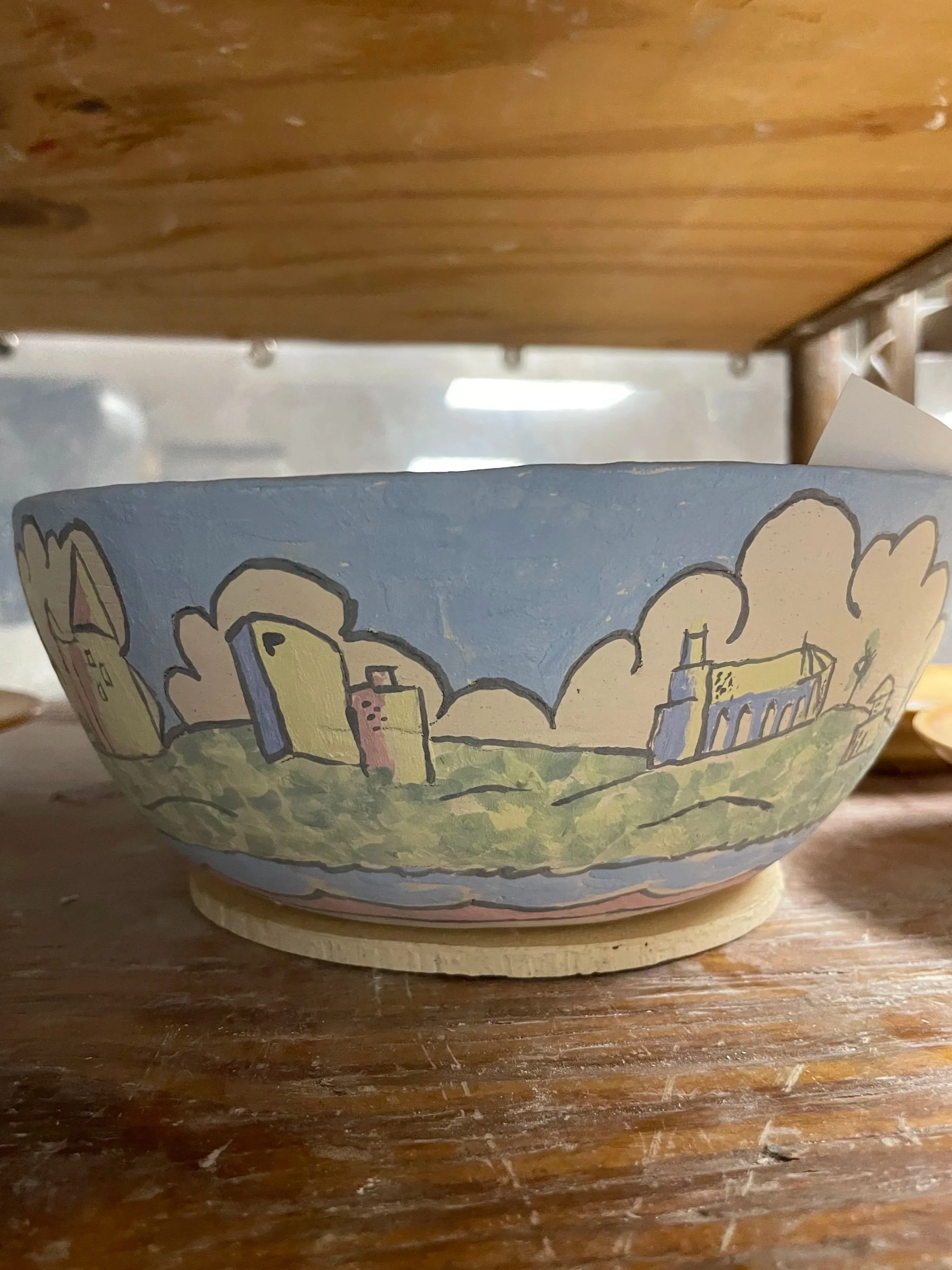A hand-painted ceramic bowl with a cityscape and cloud design, waiting to be fired in a kiln.