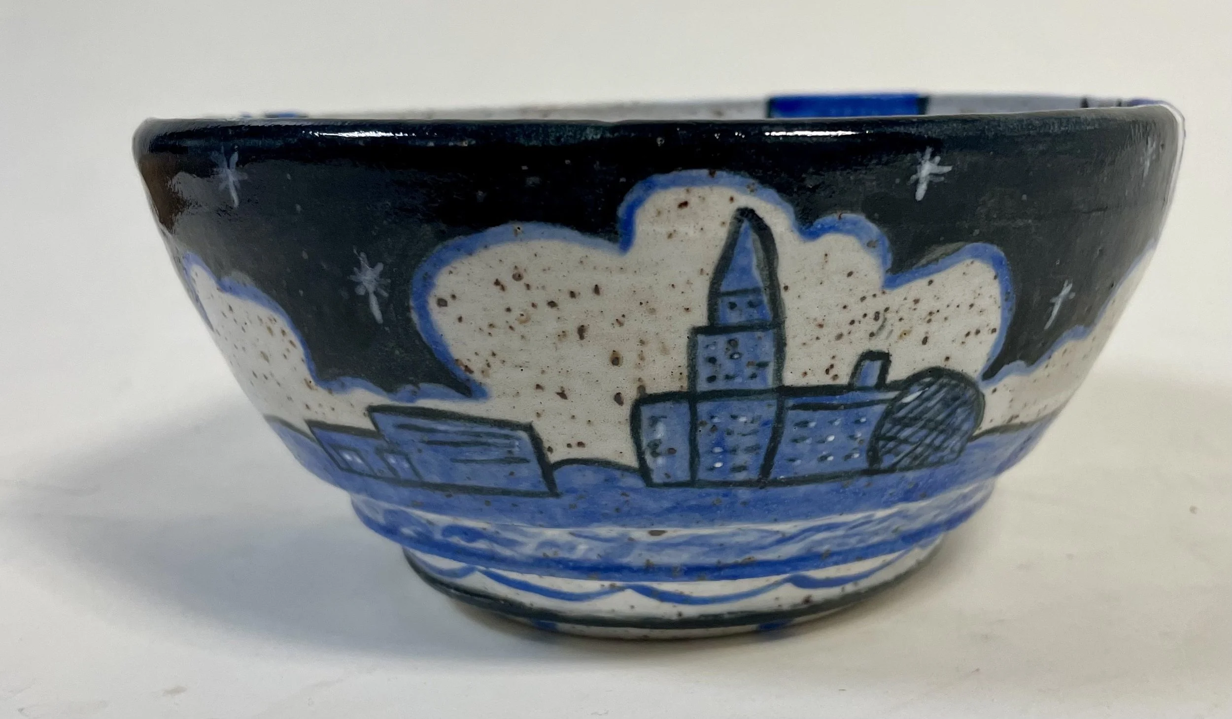 Outside of a ceramic bowl with a nautical scene featuring a lighthouse, buildings, and water, painted in blue, black, and white.