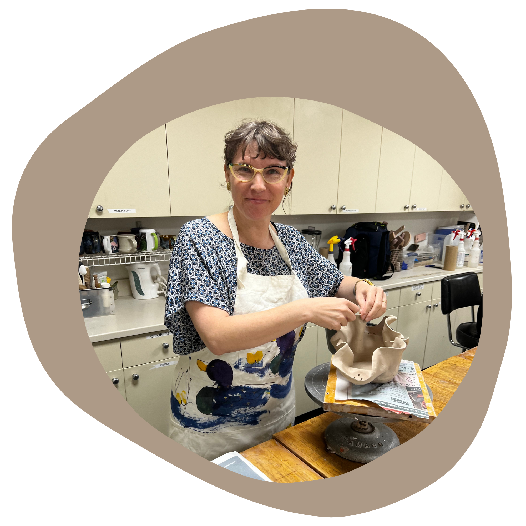 A woman wearing glasses and a patterned apron working on a clay piece in a ceramic studio, surrounded by supplies and equipment.