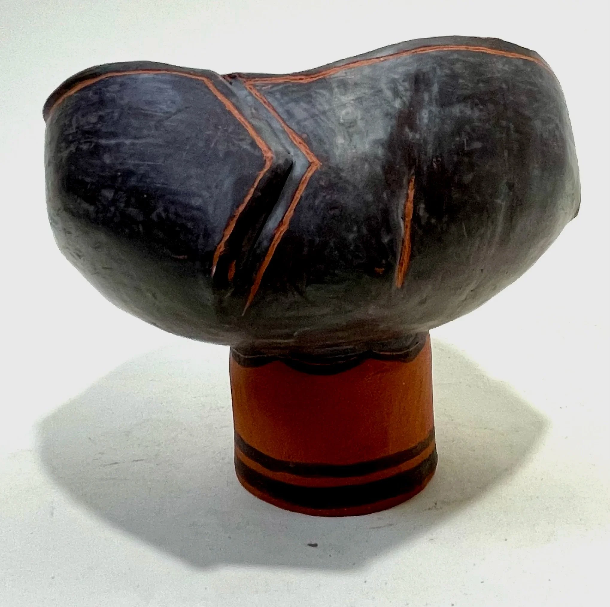 A ceramic vessel with a wide bowl on a pedestal base, glazed in dark gray and terracotta colors, before firing.