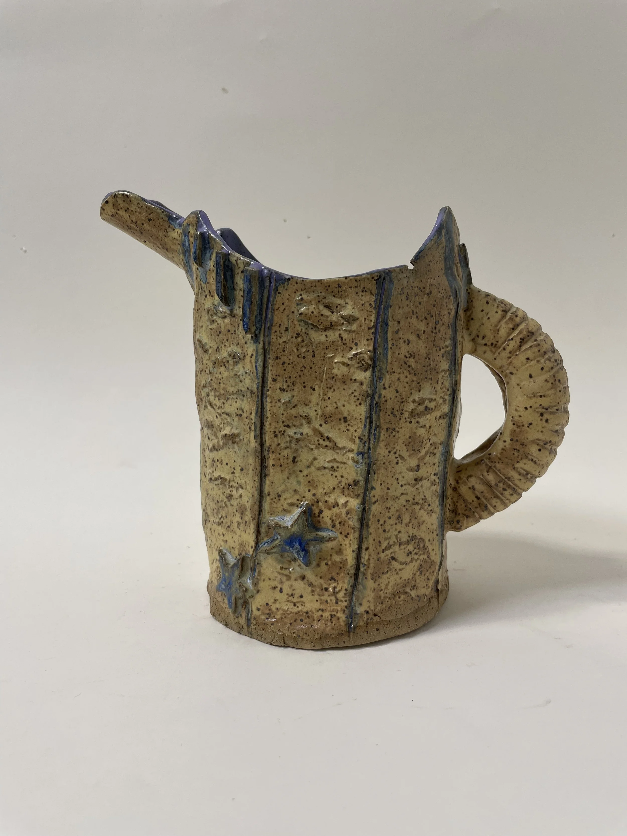 A small ceramic pitcher with a handle and a long lip, with a textured beige exterior decorated with blue stars and lines, and blue glaze inside.