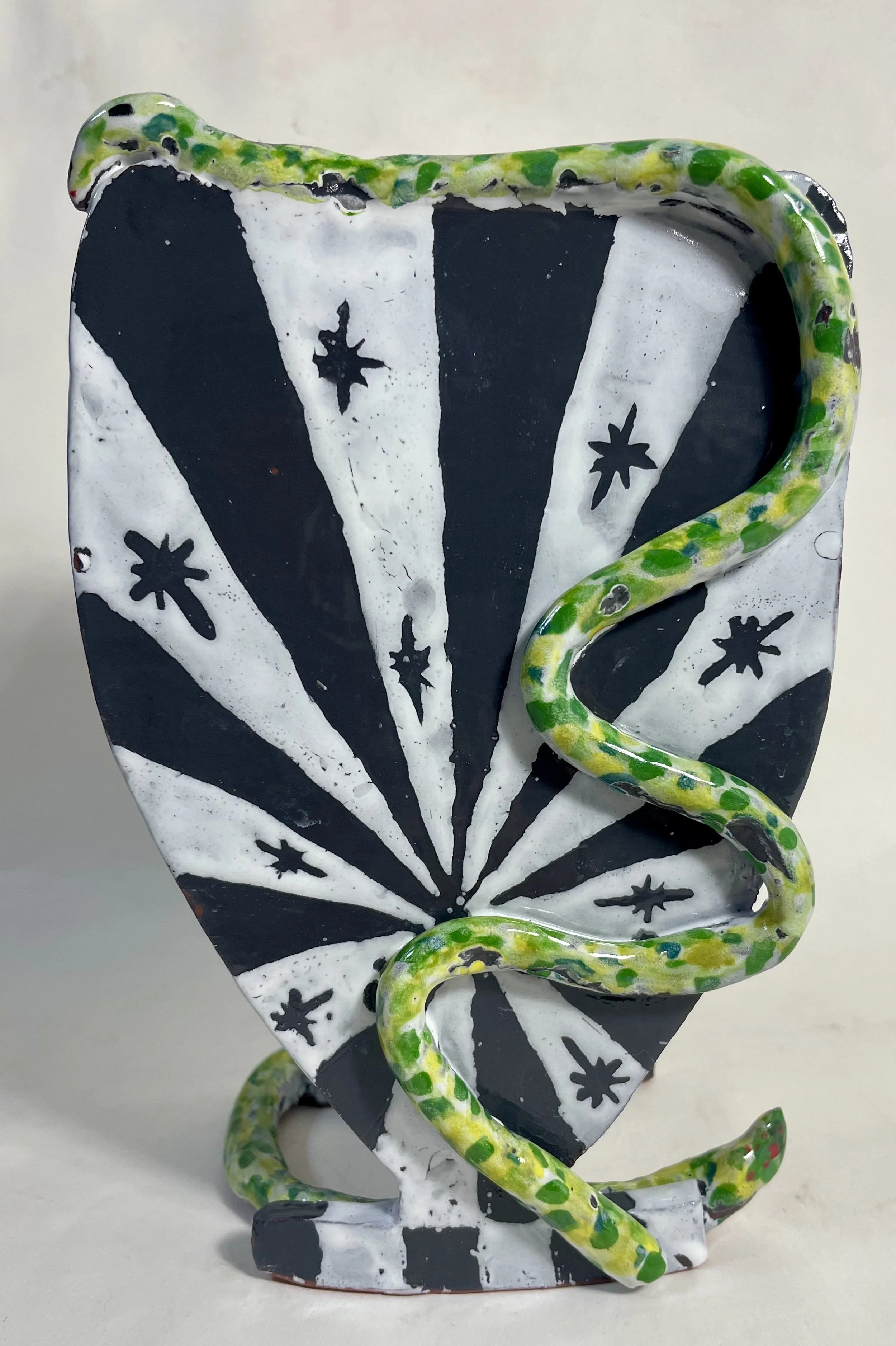 Decorative ceramic shield with black and white spiral pattern and star shapes, featuring green and yellow spotted snakes coiled around it.