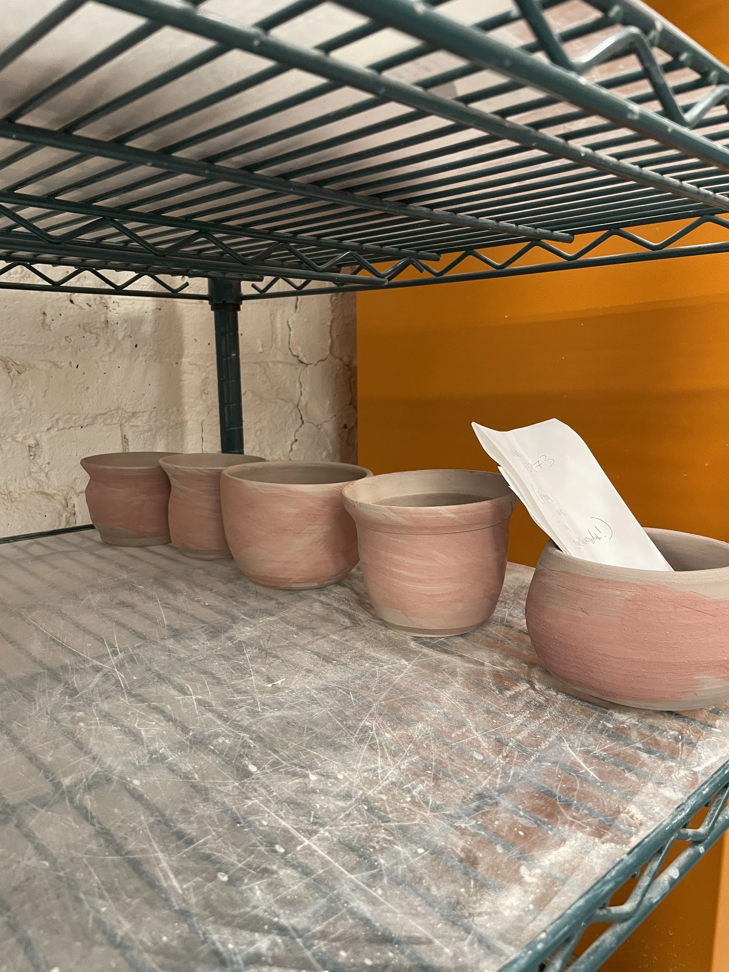 Five unfired clay pots on a wire shelving unit against a white brick wall and orange background.