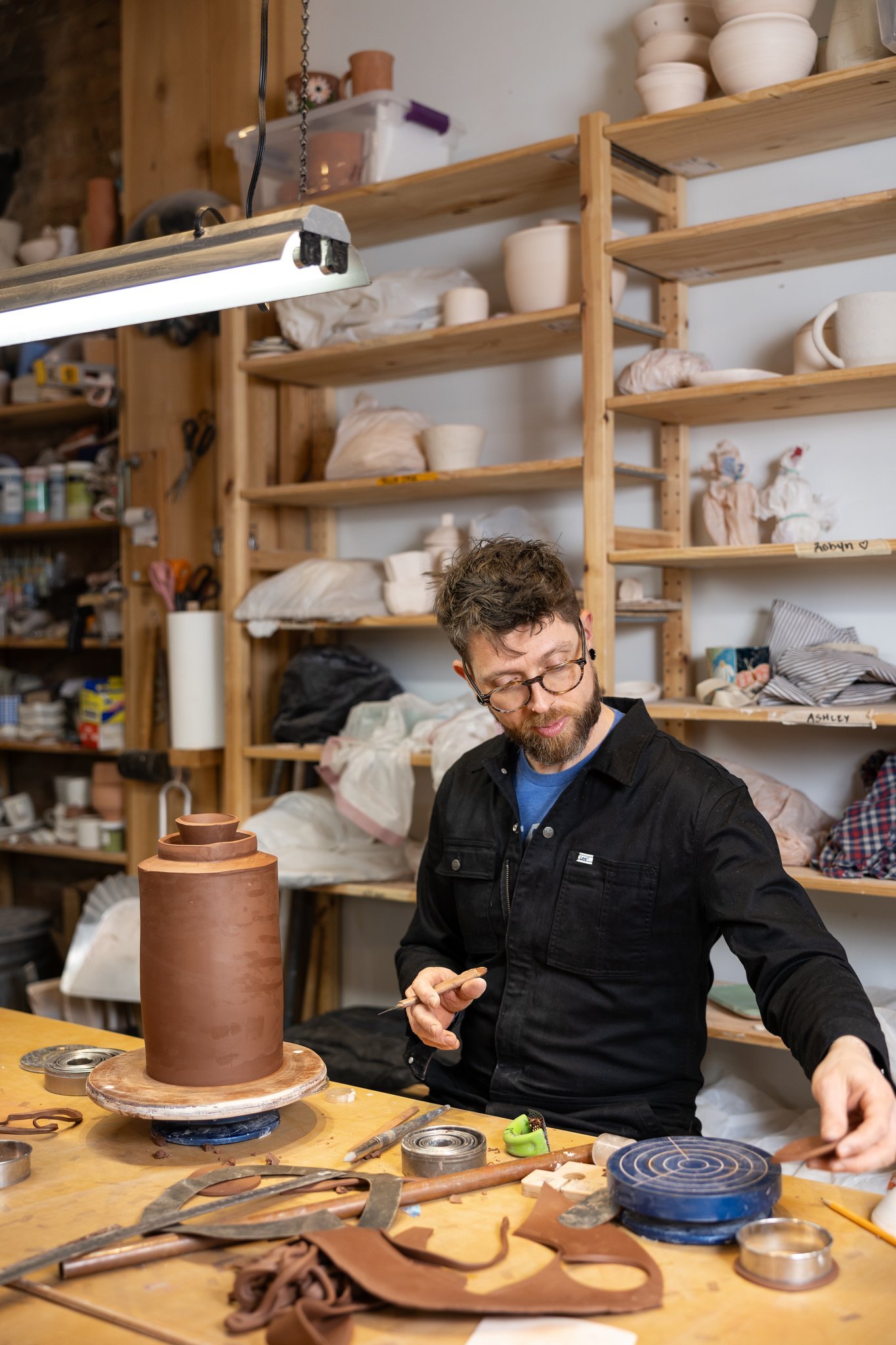 A man working at a pottery studio, shaping a large clay vase on a rotating wheel with various pottery tools and materials on the work table.