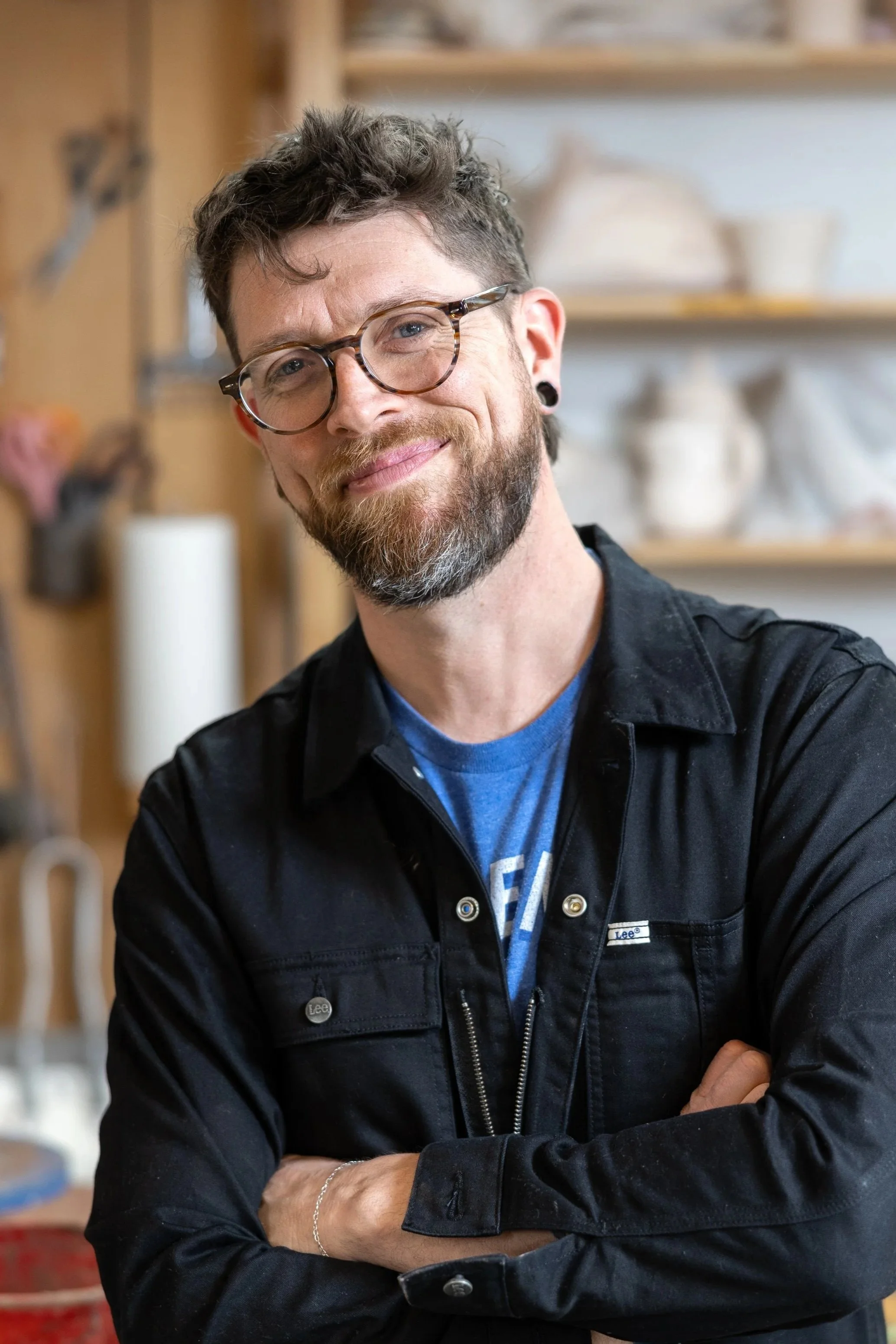 A man with glasses and a beard smiling with his arms crossed, standing in a woodworking or pottery studio with shelves and supplies in the background.