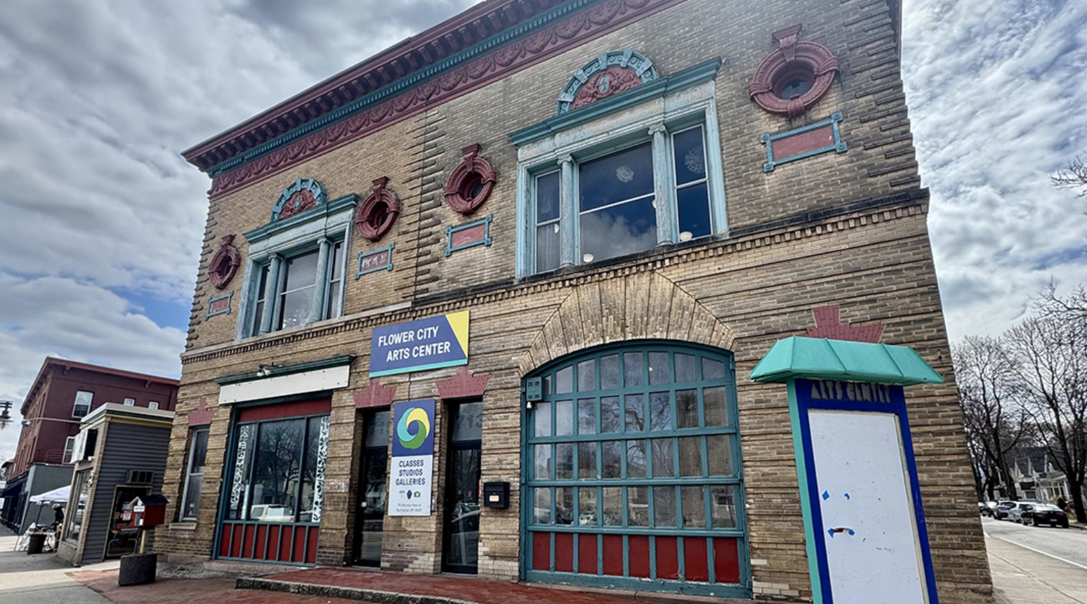 Brick building with blue and red decorative trim, large arched window, and signage for Flower City Arts Center, with a sidewalk and parking lot in front.