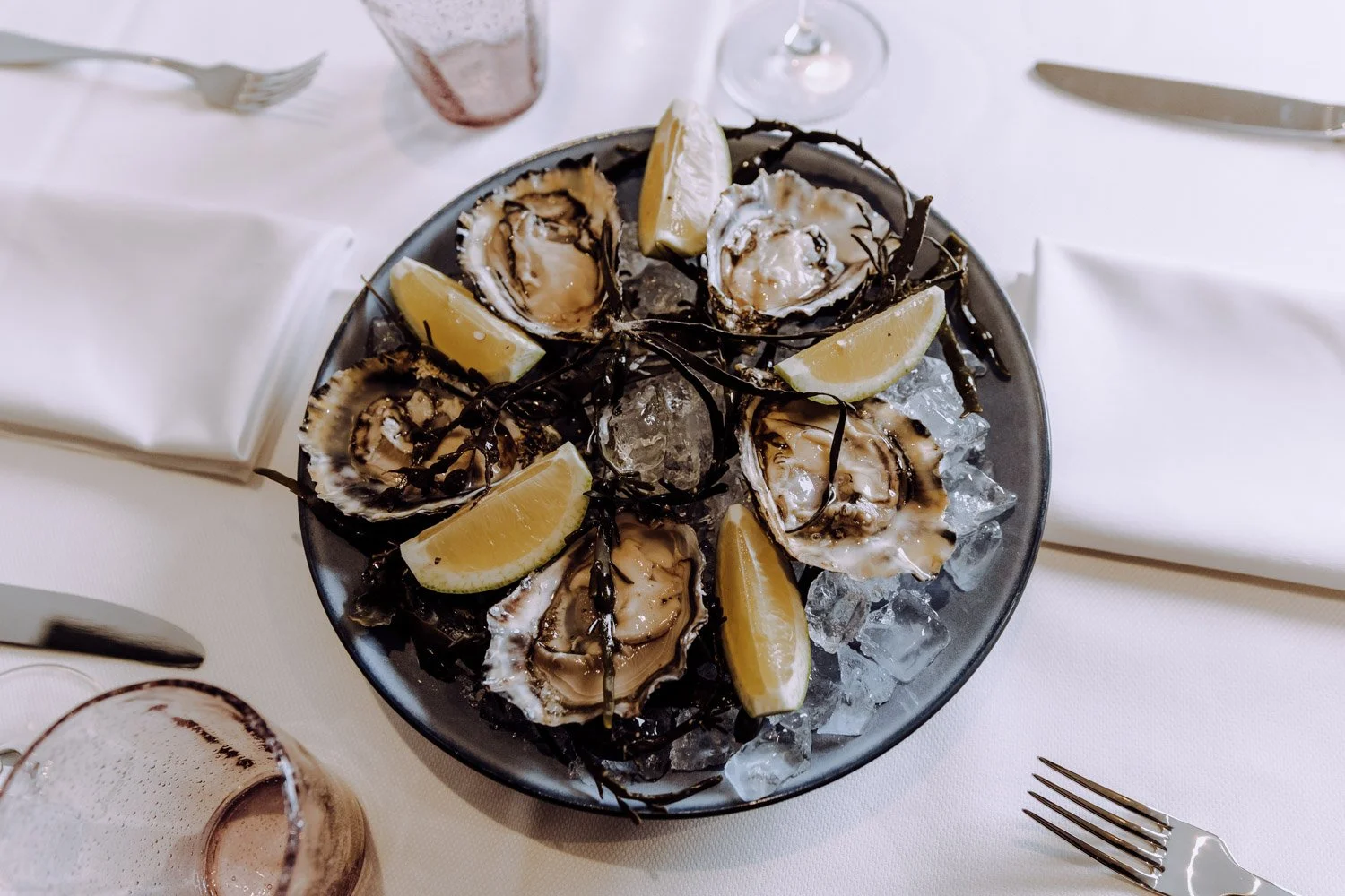 A plate with fresh oysters on ice, garnished with lemon wedges and chocolate drizzle, arranged on a table set for dining.