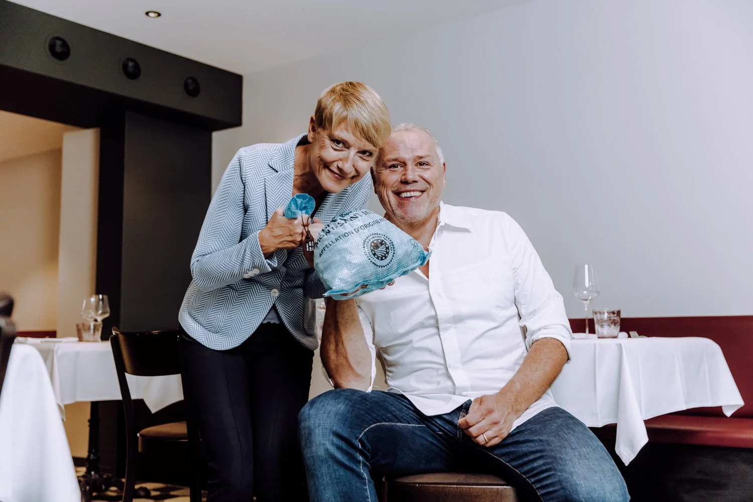 Lächelnder älterer Mann im weißen Hemd sitzt an einem Tisch in einem Restaurant, während eine ältere Frau im blauen Blazer neben ihm steht und ihm ein Geschenk reicht.