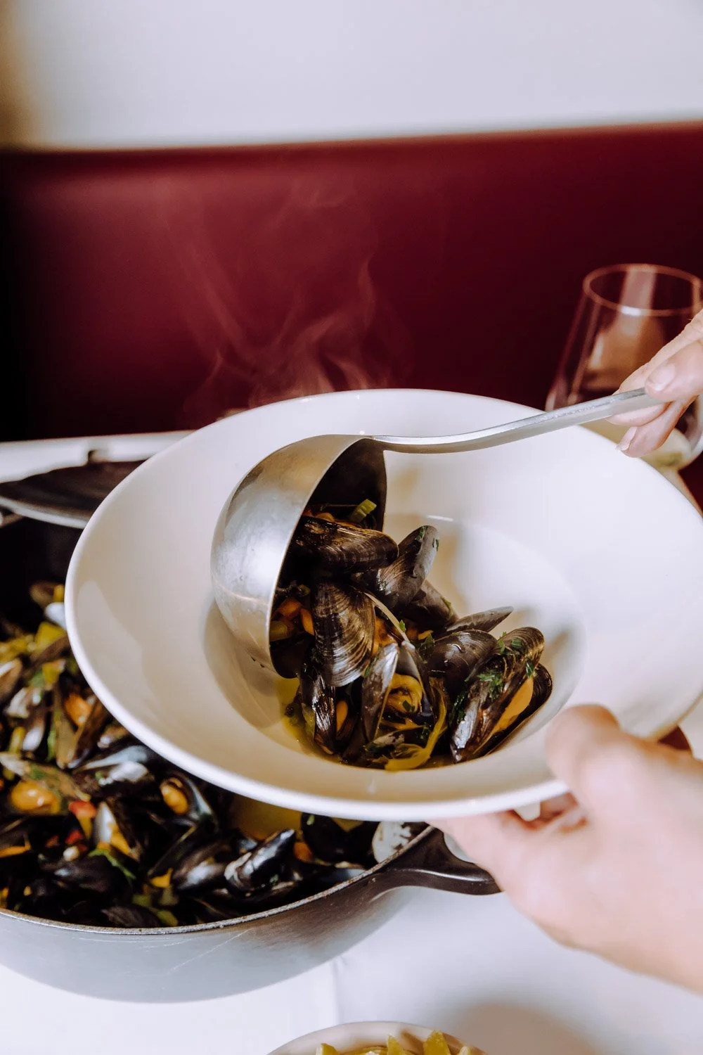 Eine Person serviert mit einer Zange Muscheln aus einem großen Topf, auf einem weißen Teller. Es riecht nach frisch gekochten Meeresfrüchten.