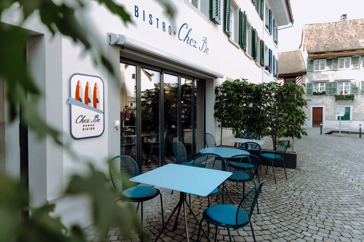 Außenansicht eines Restaurants namens 'Chez Be Bistro' mit blauen Tischen und Stühlen, an einer gepflasterten Straße in einem europäischen Ort, mit historischen Gebäuden im Hintergrund.