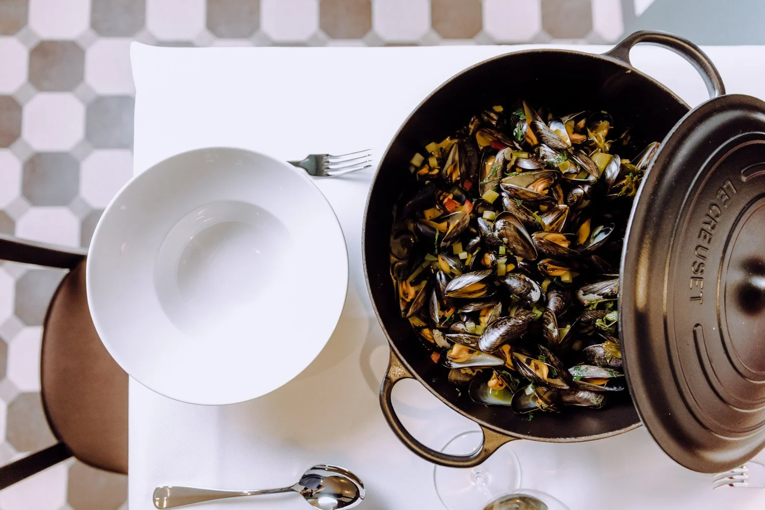 Schüssel mit Muscheln und Gemüse, daneben leere weiße Schale, Gabel, Löffel und Weinglas auf weißem Tisch.