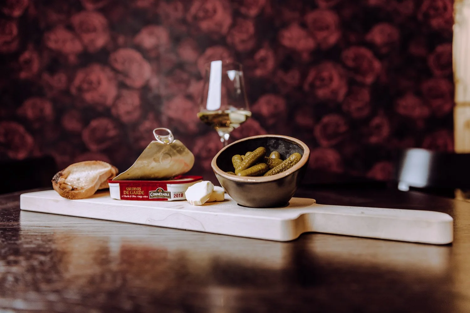 Käse, Sardinen, Salat, Brot und ein Glas Weißwein auf einem Holztisch.