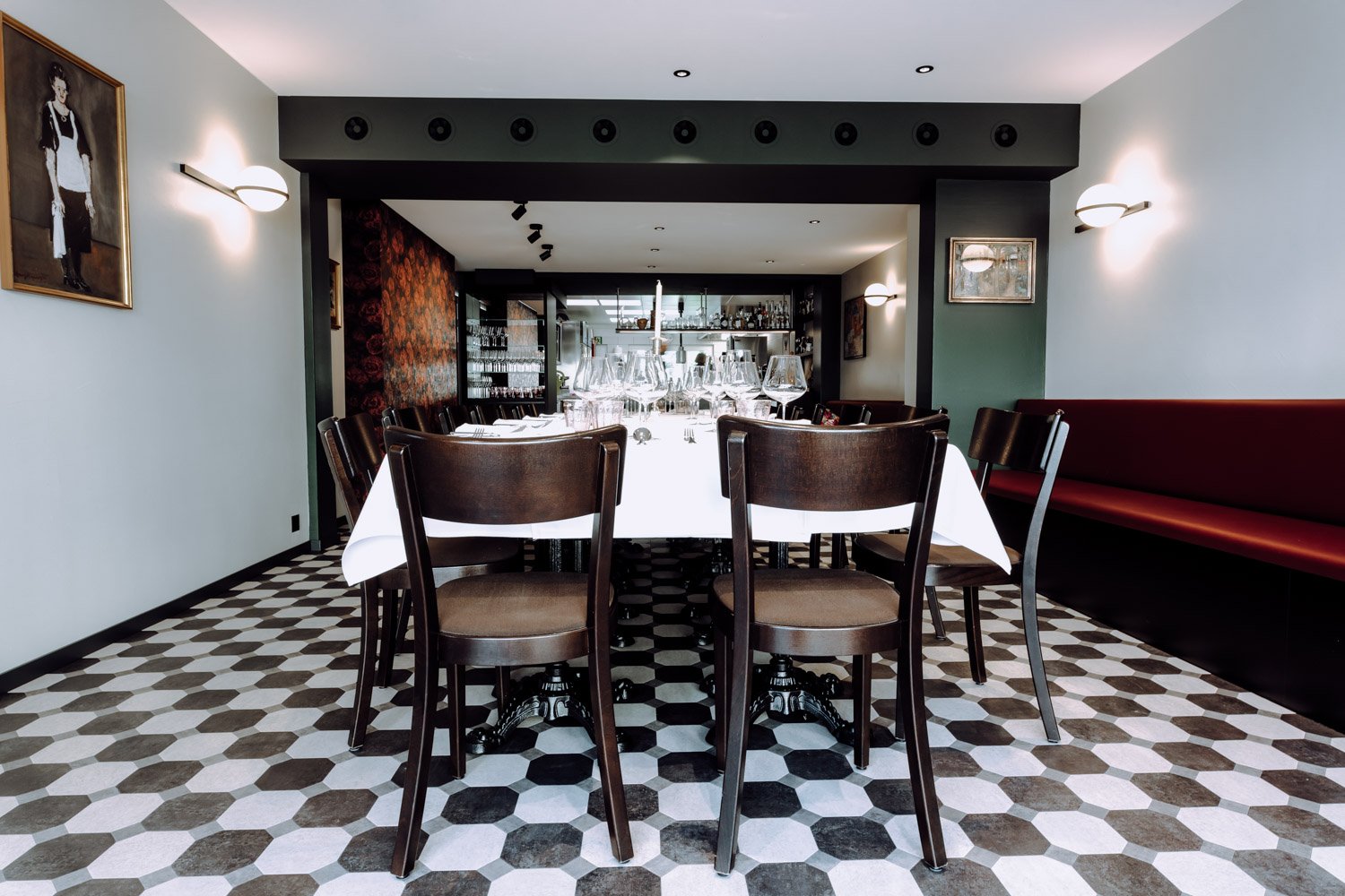 Ein elegant gedeckter Esstisch im Restaurant mit mehreren Weingläsern, dunkle Holztische und Stühle, Wanddekorationen, weiße Wände, gemusterter Boden, im Hintergrund die Bar.